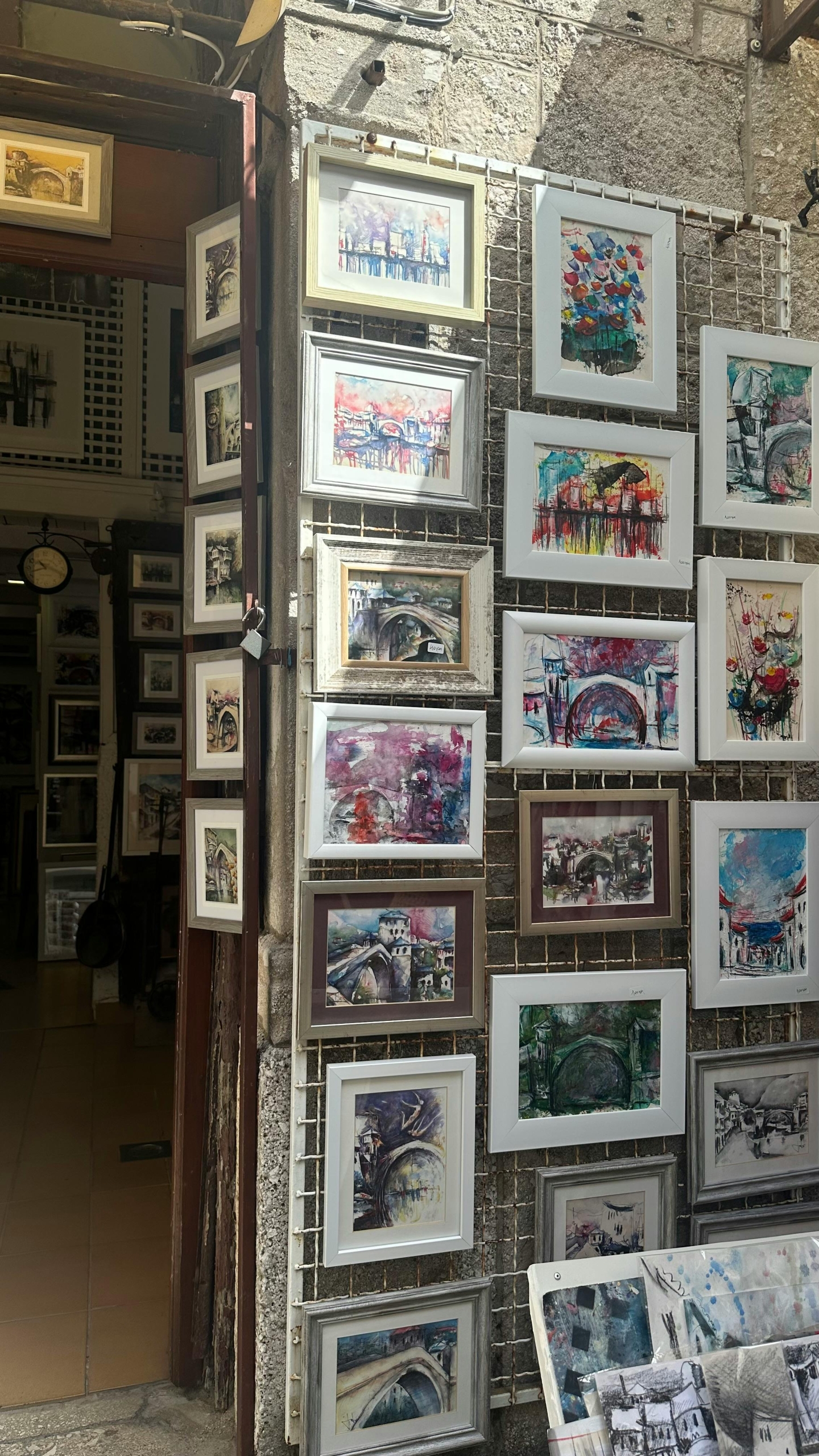 A display of framed watercolor and acrylic paintings hanging on a white wire grid rack outside a shop or market stall.