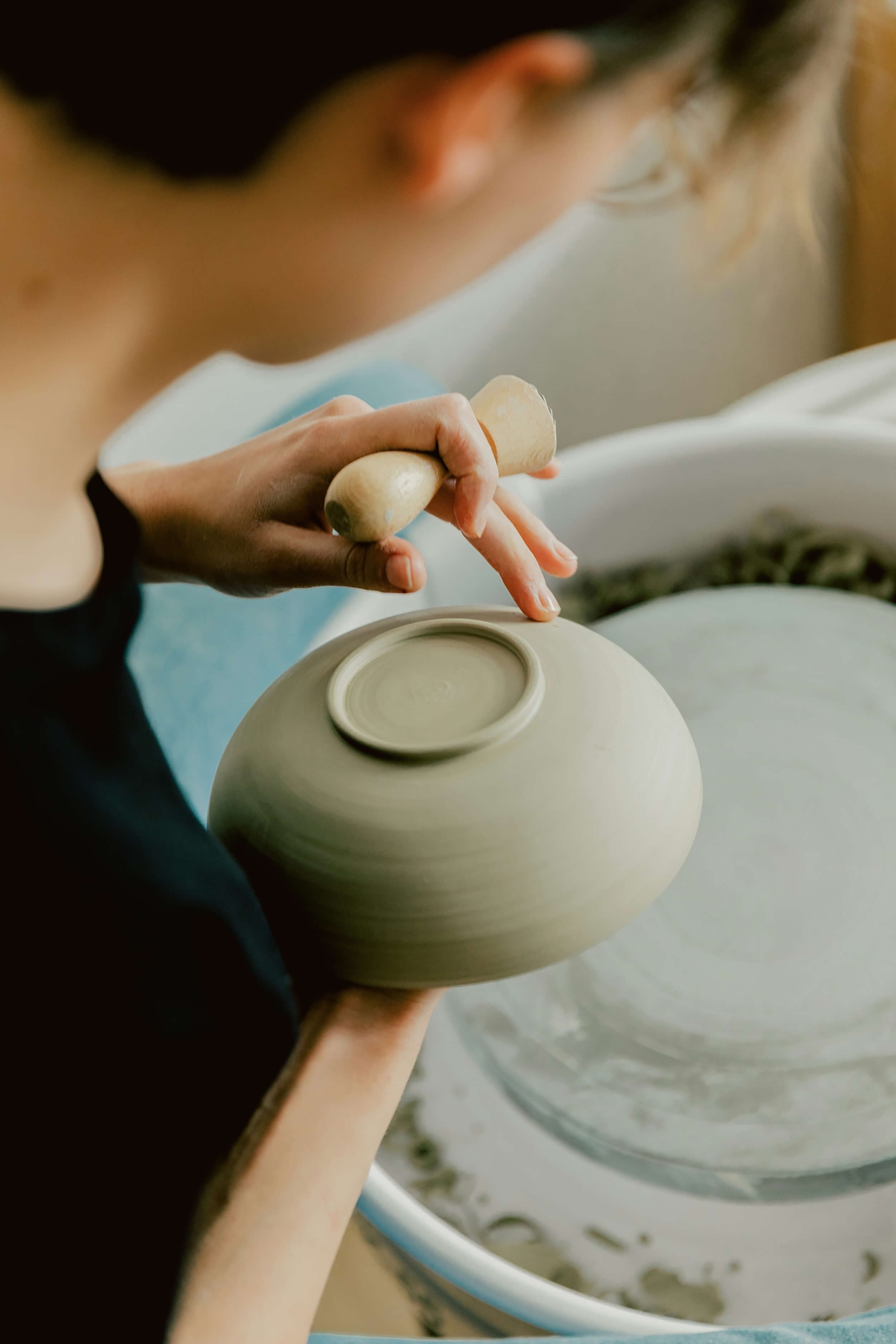 A close-up of a potter using a wooden tool to trim and shape the base of a clay bowl while it spins on a pottery wheel.