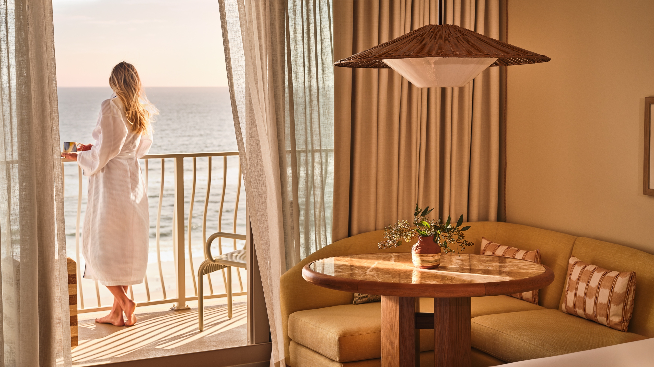 A woman in a white robe stands on a hotel balcony with a coffee mug, looking out at the ocean.