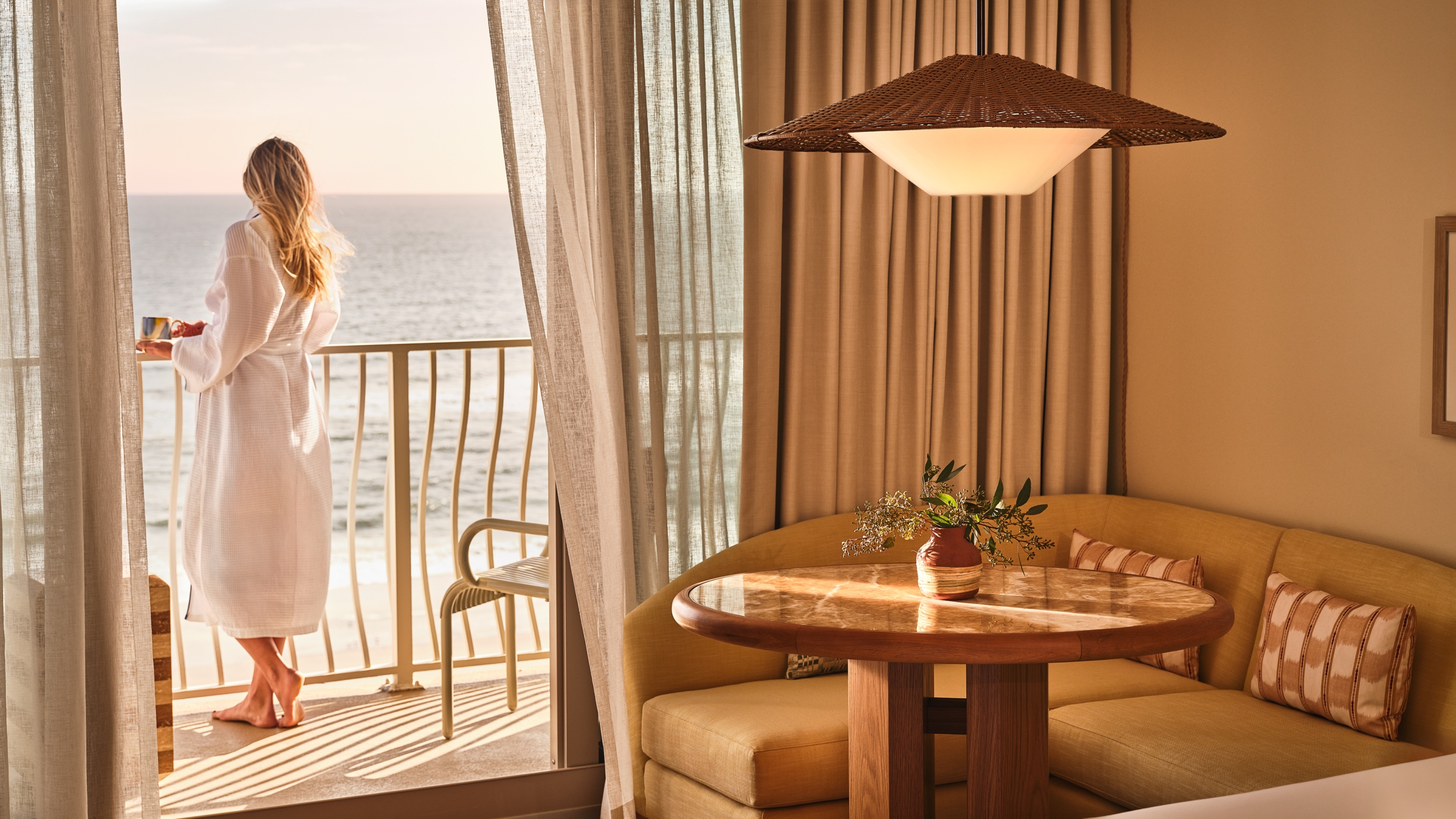 A woman in a white robe standing on a balcony with a coffee mug, looking out at the ocean, viewed from inside a hotel room with a dining area.