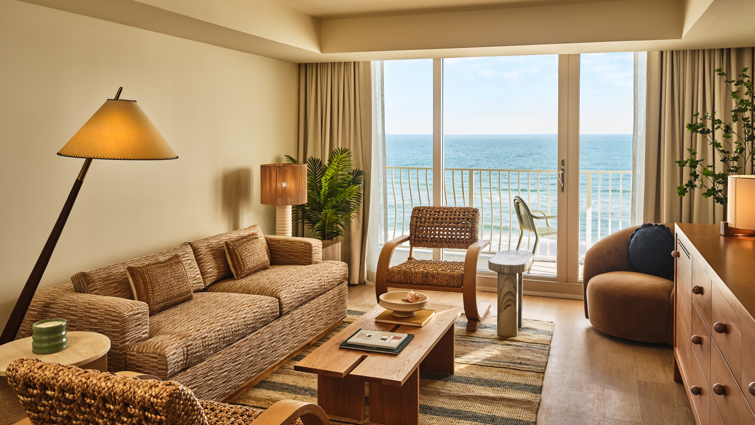 A bright hotel room living area featuring a beige sofa, wicker armchairs, and a wooden coffee table, with sliding glass doors opening to an ocean view balcony.