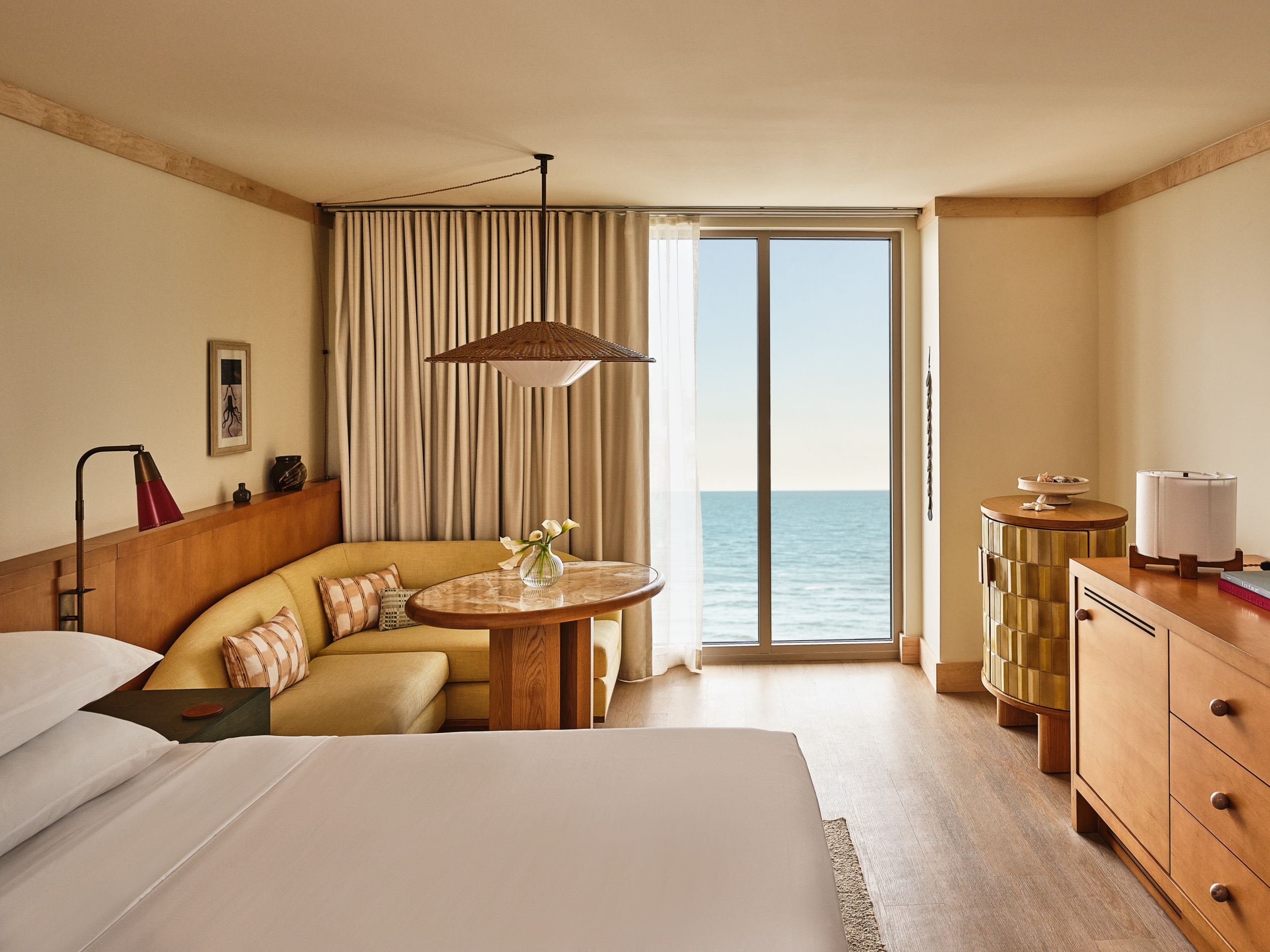 A modern hotel room featuring a bed, a yellow corner sofa, a round table, and a wooden dresser, with a large window overlooking the ocean.