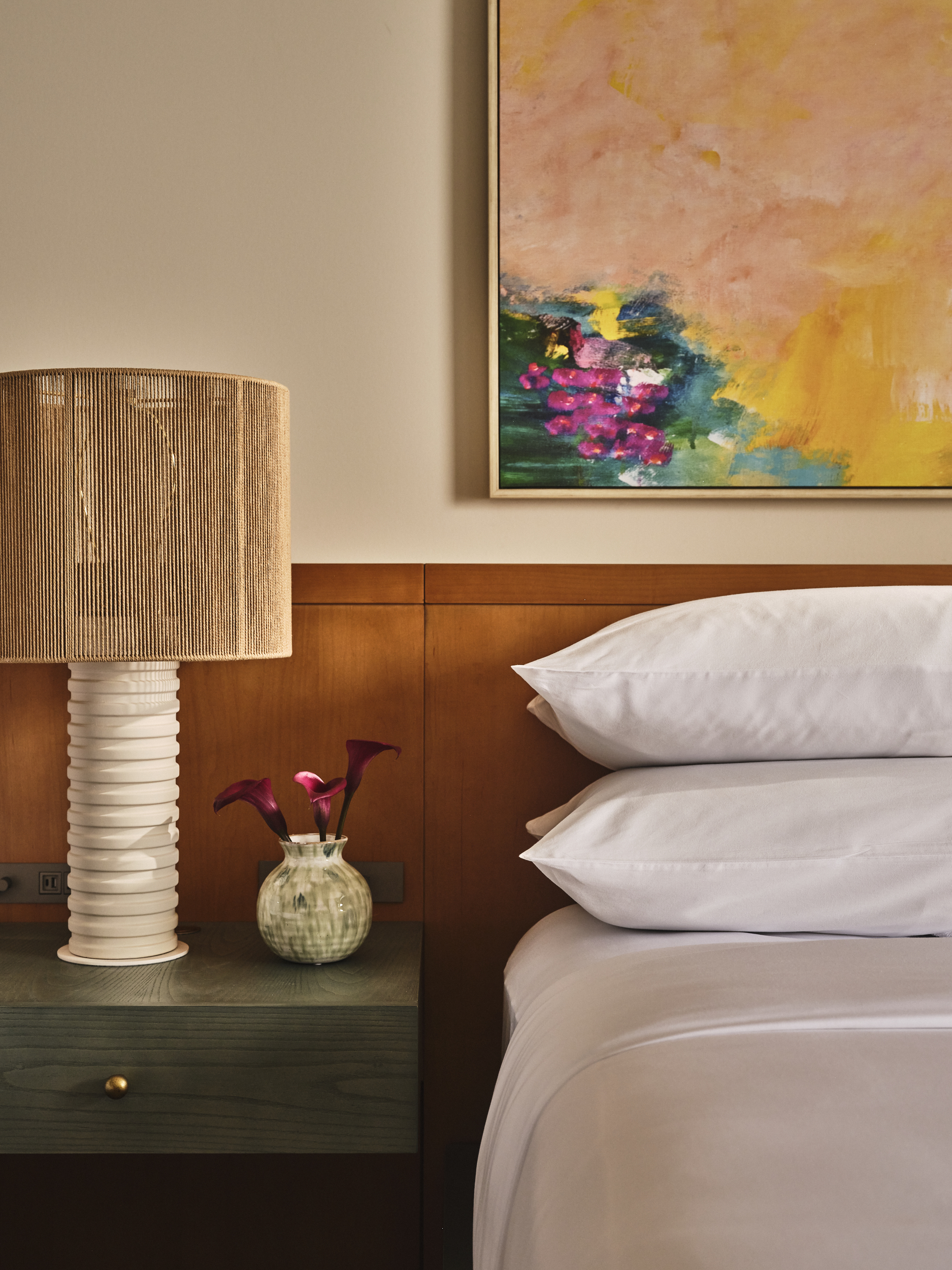 Close-up of a teal bedside table with a tall textured lamp, a small vase of purple flowers, and white pillows.