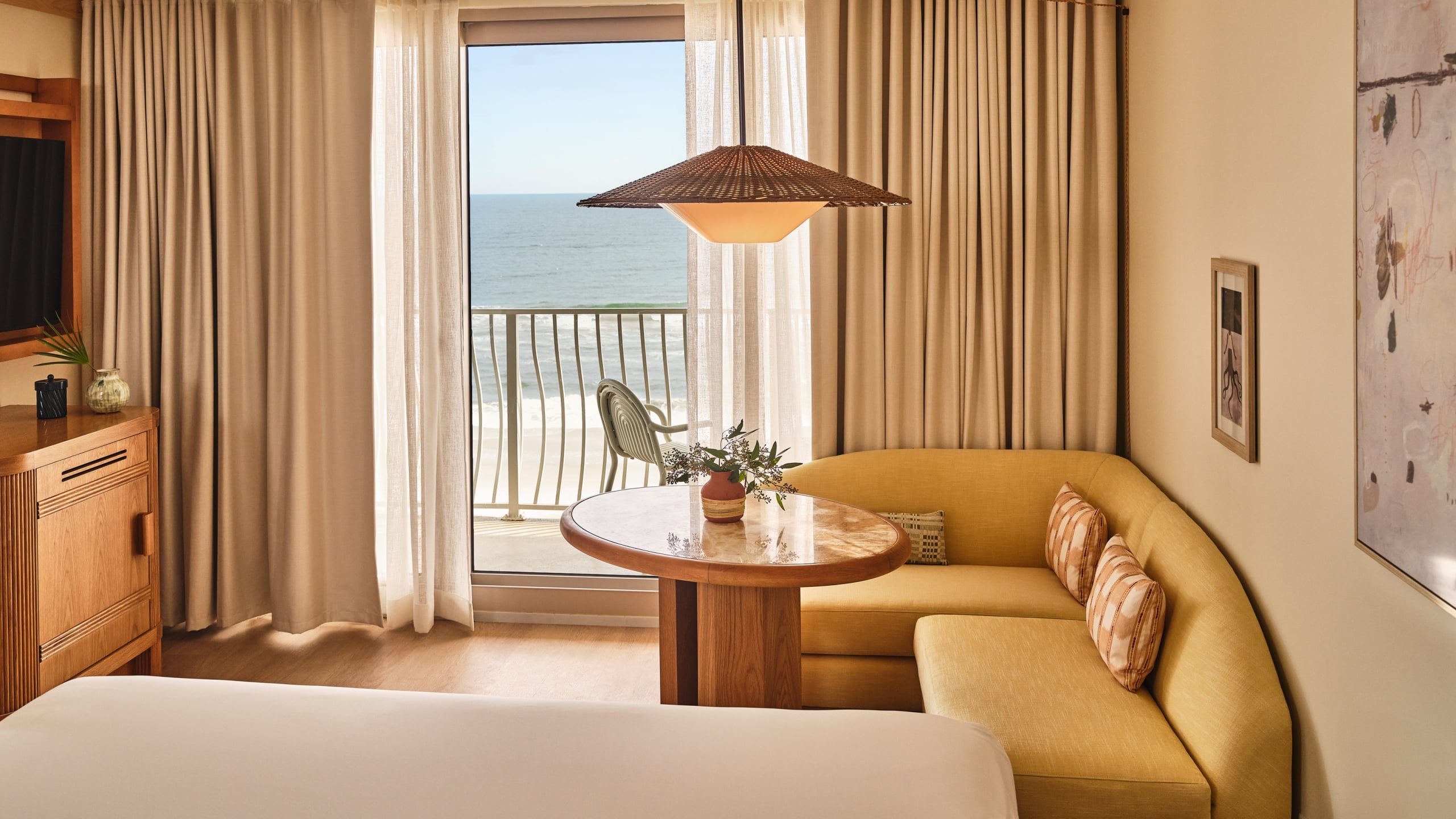A hotel room corner with a yellow curved sofa, a marble table, and a balcony view of the ocean waves.