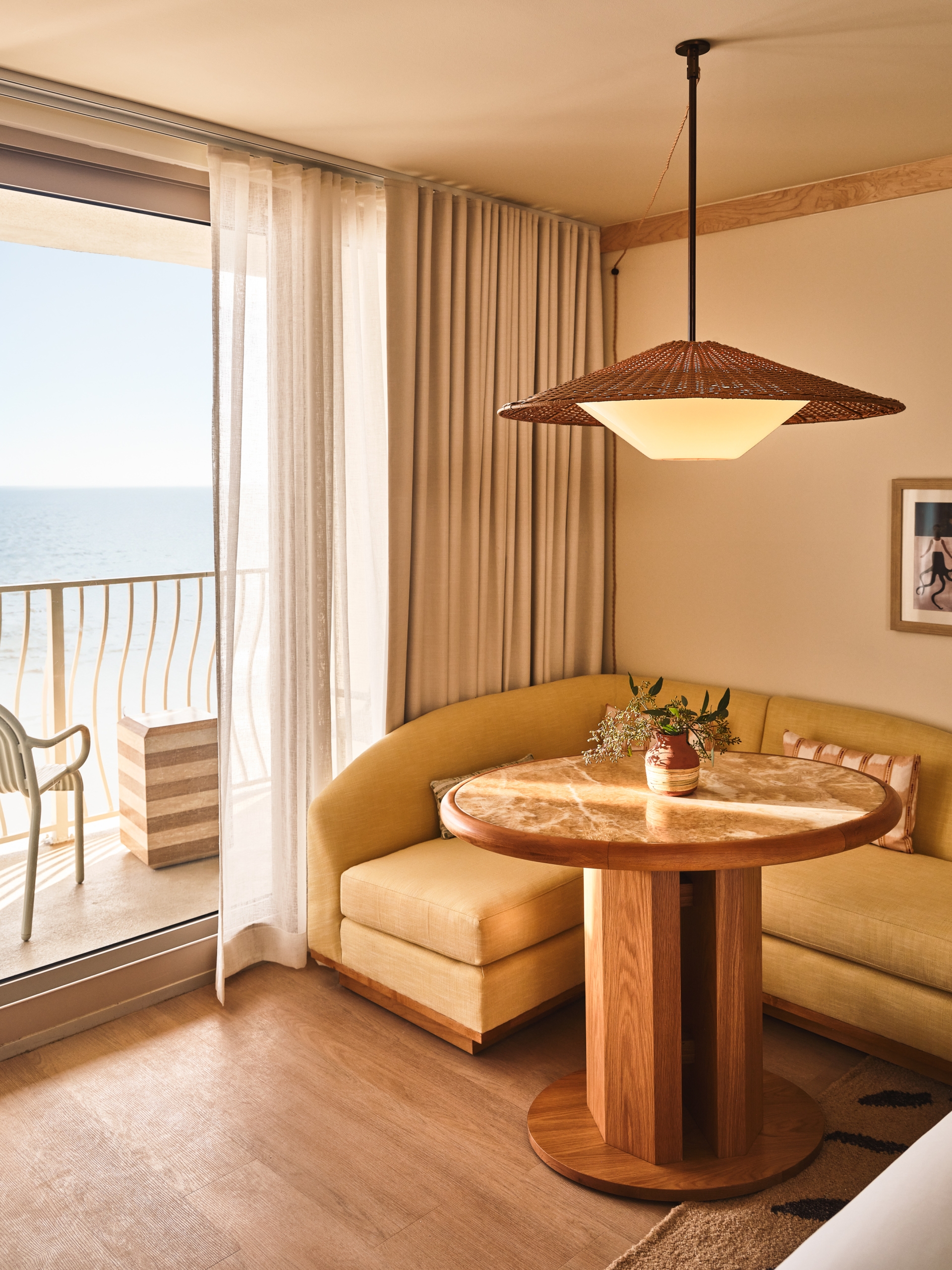 Vertical view of a hotel seating area with a yellow sofa, marble table, and a woven pendant light overlooking the sea.