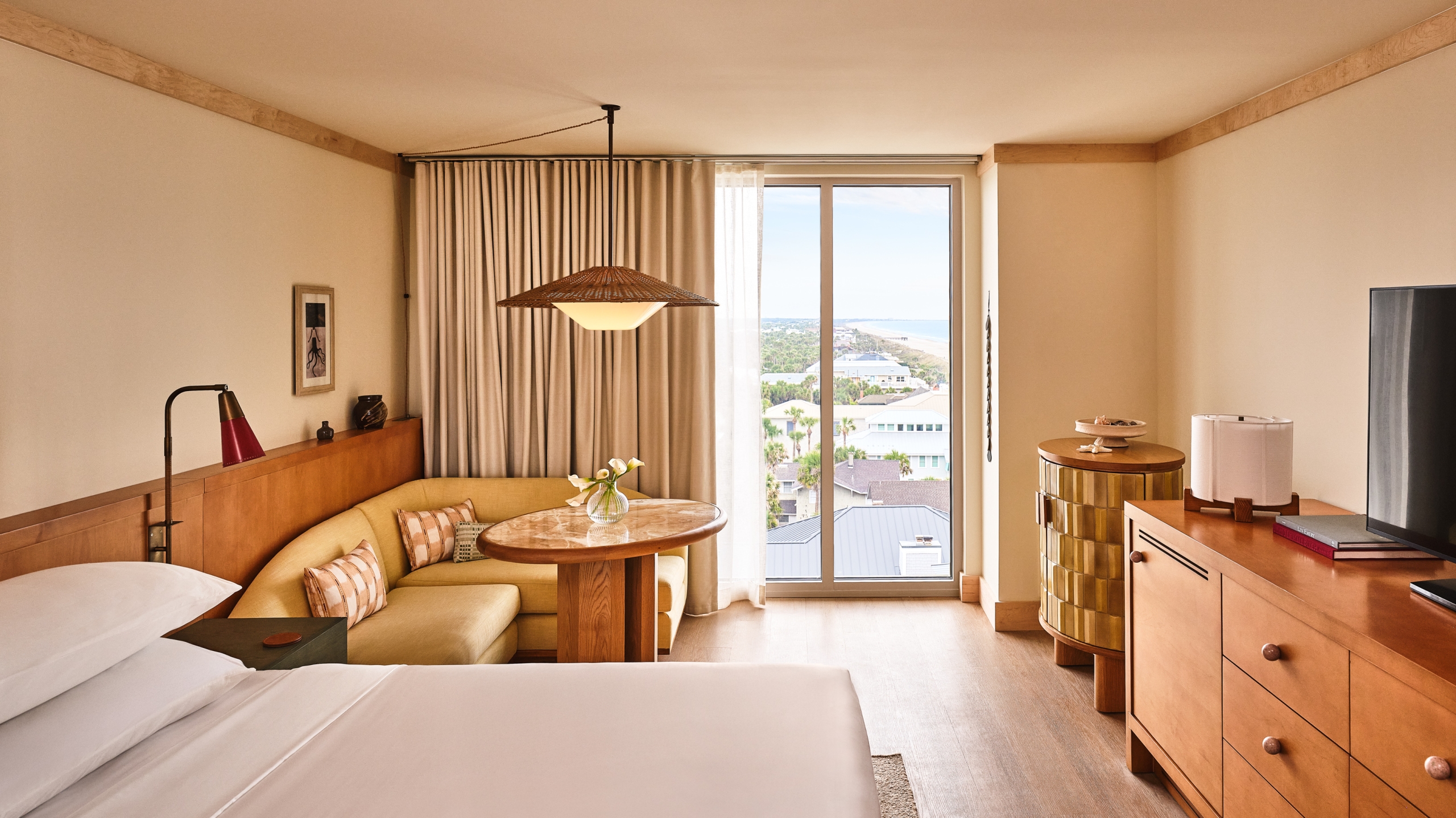 View from a hotel bed showing a wooden dresser, a sitting area, and a window facing the coastline and distant beach.