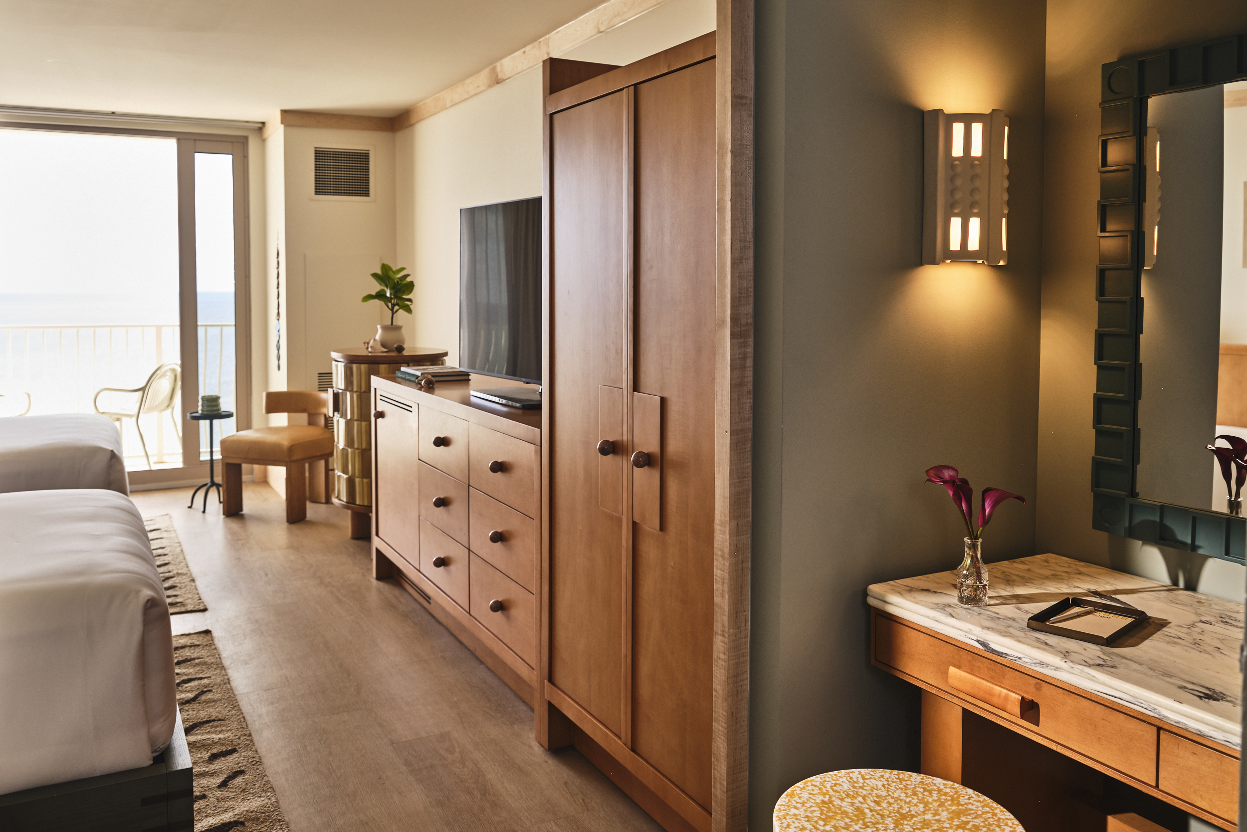 A hotel room view looking past a wooden dresser and wardrobe toward a bright balcony and a marble vanity nook.