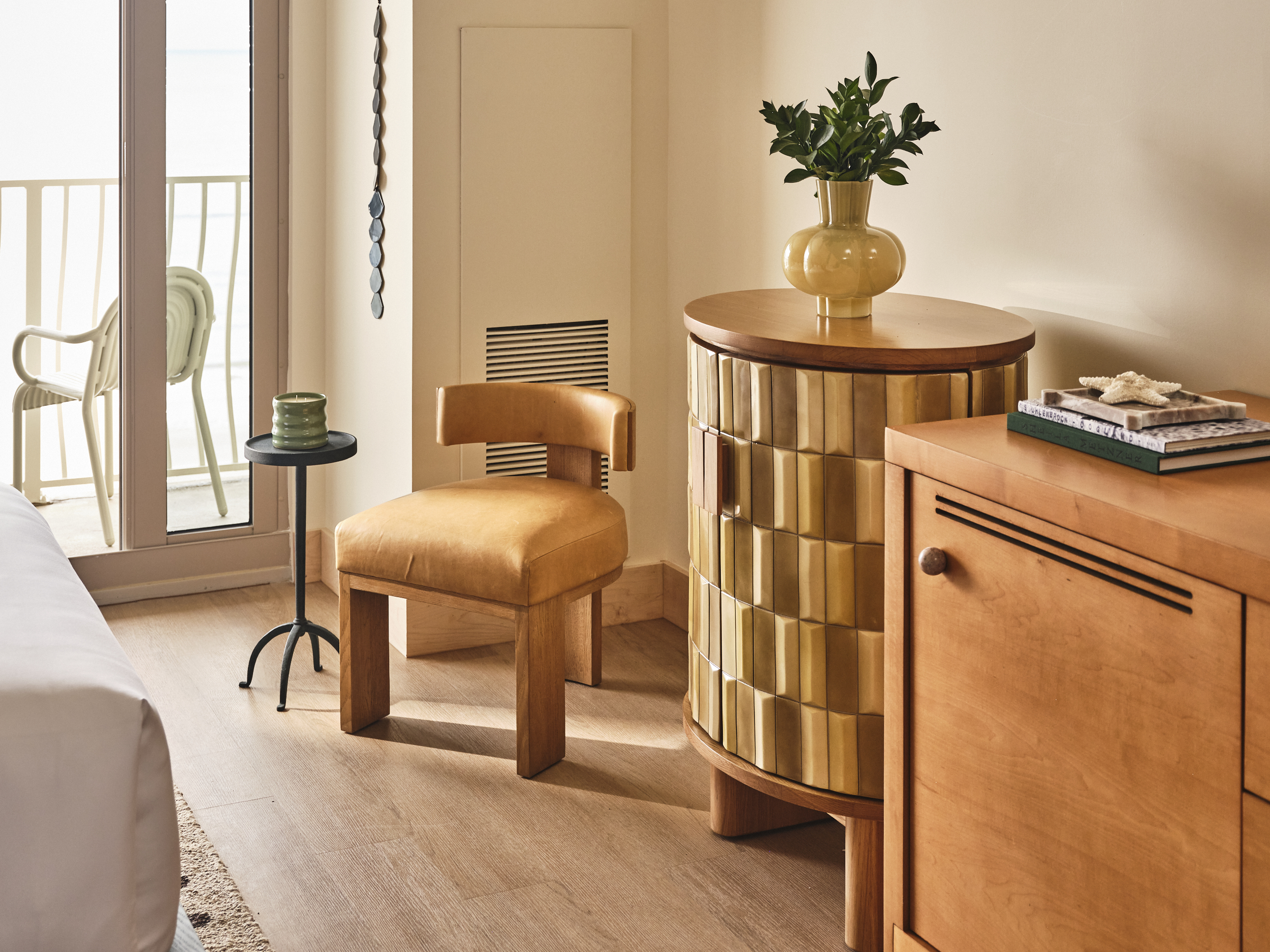 Dune_House_Room_Queen_Balcony_Seating.jpg A stylish hotel room corner featuring a tan leather chair, a gold cylindrical cabinet, and a balcony with an ocean view.