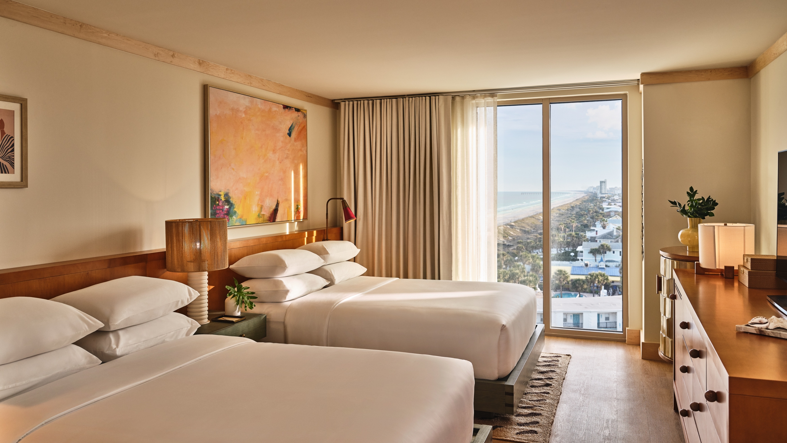 A hotel room with two white queen beds, a large abstract painting, and floor-to-ceiling windows overlooking the coast.