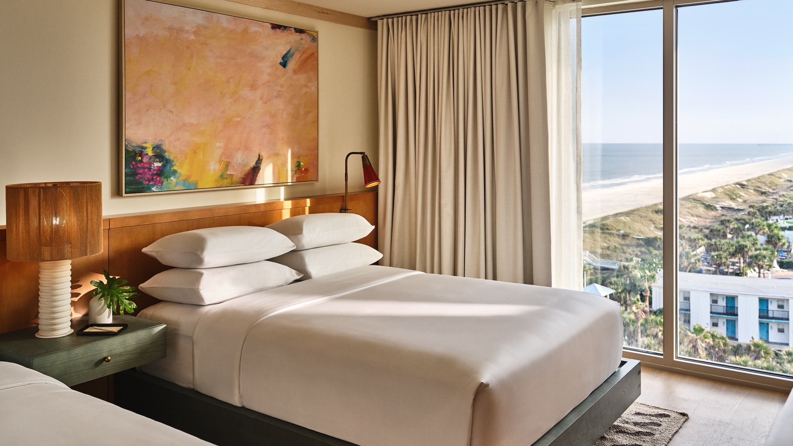 A bright hotel bedroom with a large abstract painting over the bed and a wide window view of a sandy beach and ocean.