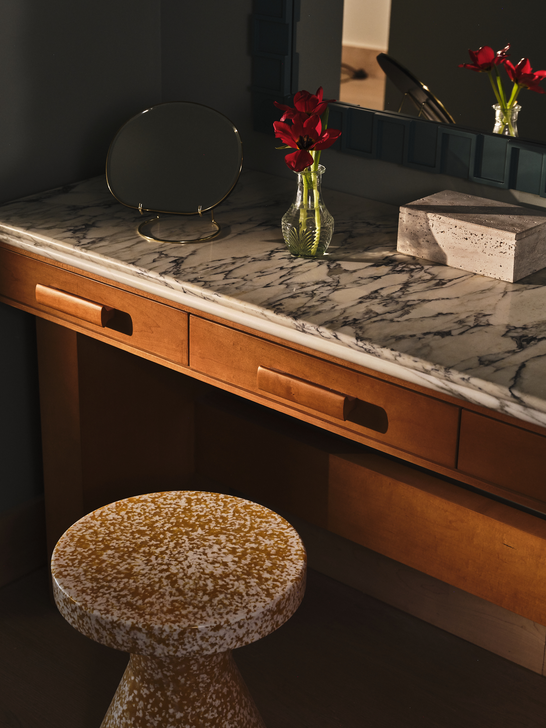 Close-up of a marble-top vanity with a terrazzo stool, a small vase of red tulips, and a gold-rimmed mirror.