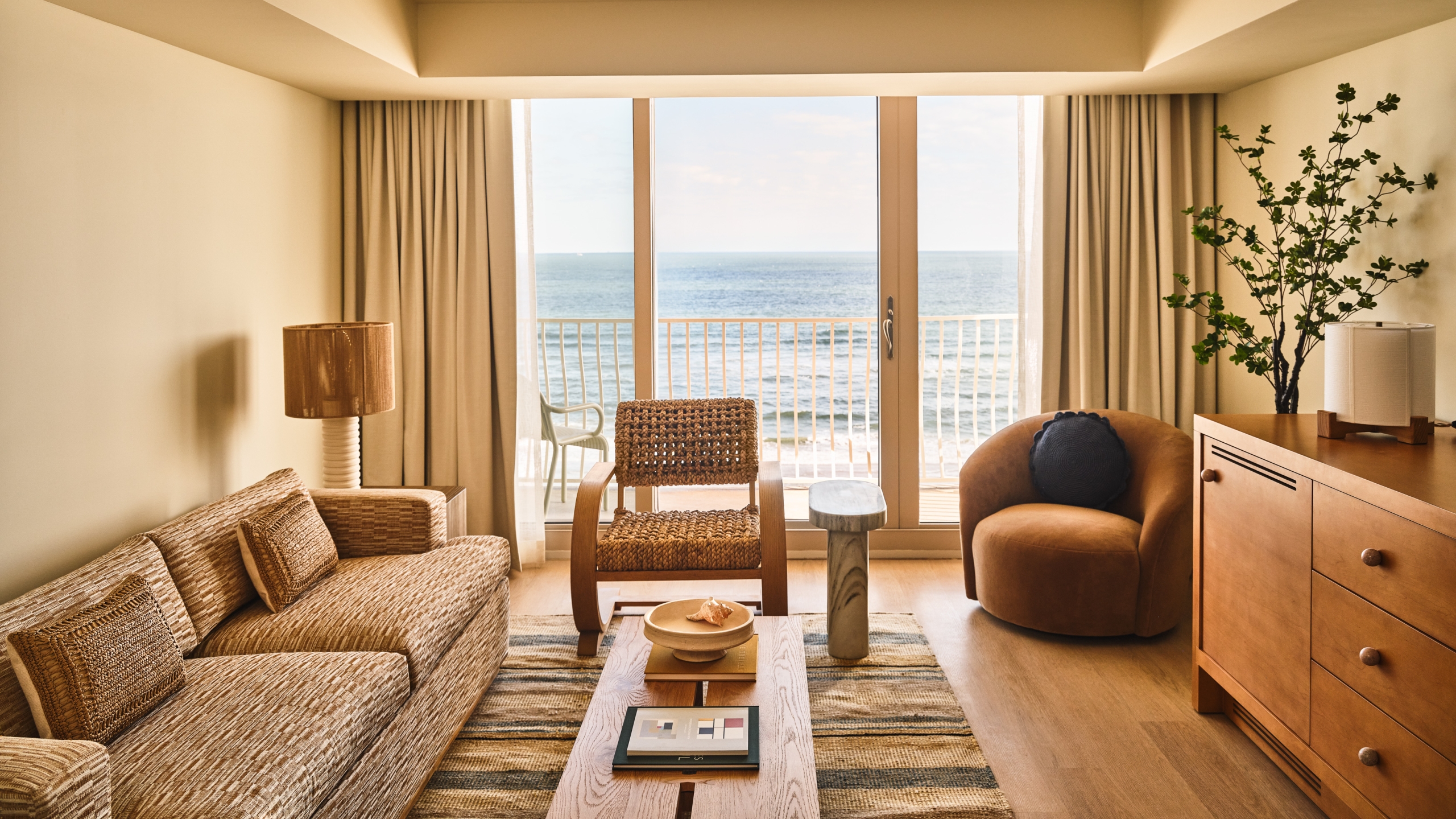 A sunlit hotel living room with a textured tan sofa, mid-century modern chairs, and a sliding glass door to the ocean.