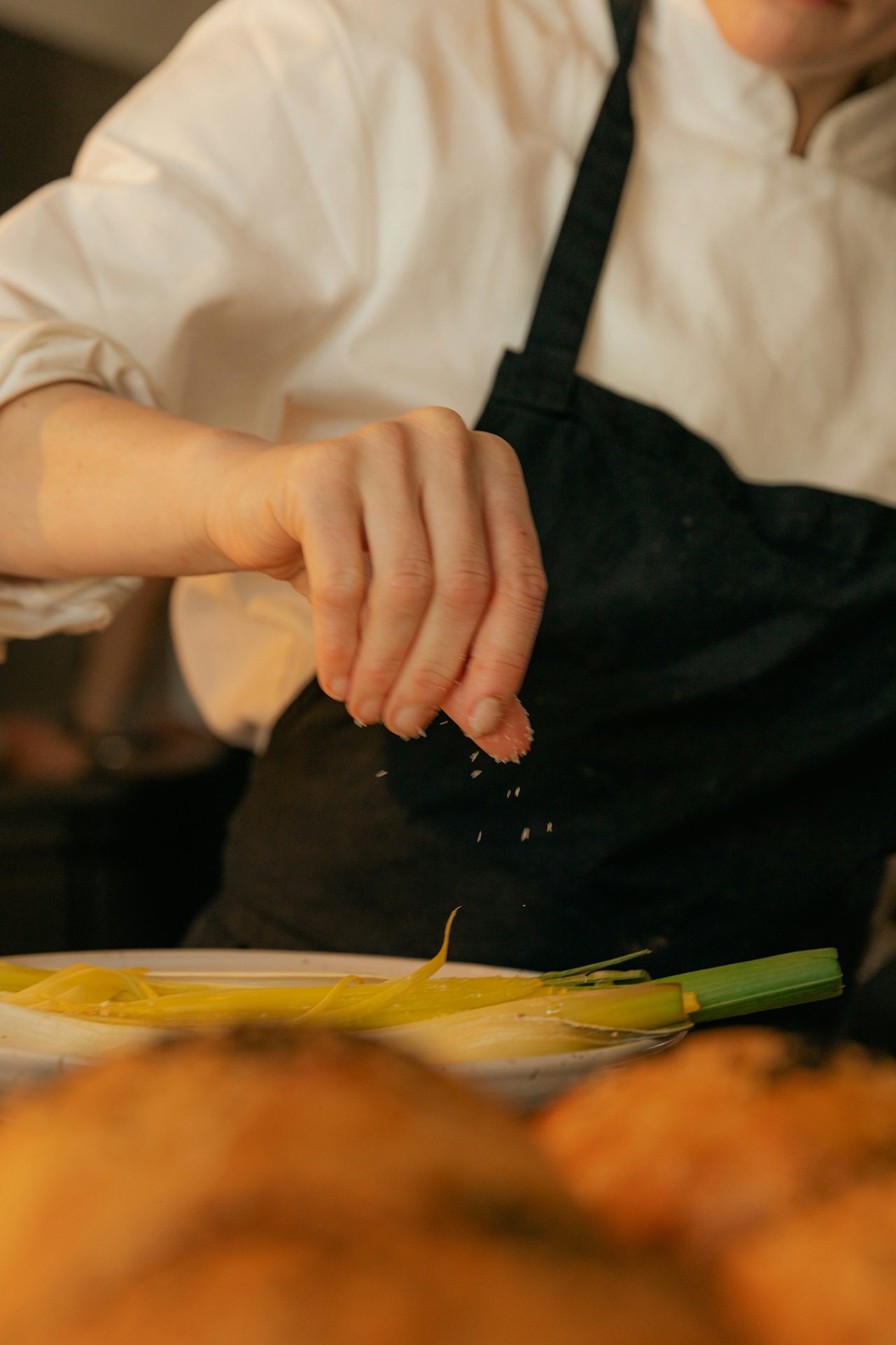 A close-up of a chef's hand sprinkling seasoning onto a plate of cooked leeks, wearing a black apron over a white chef's coat.