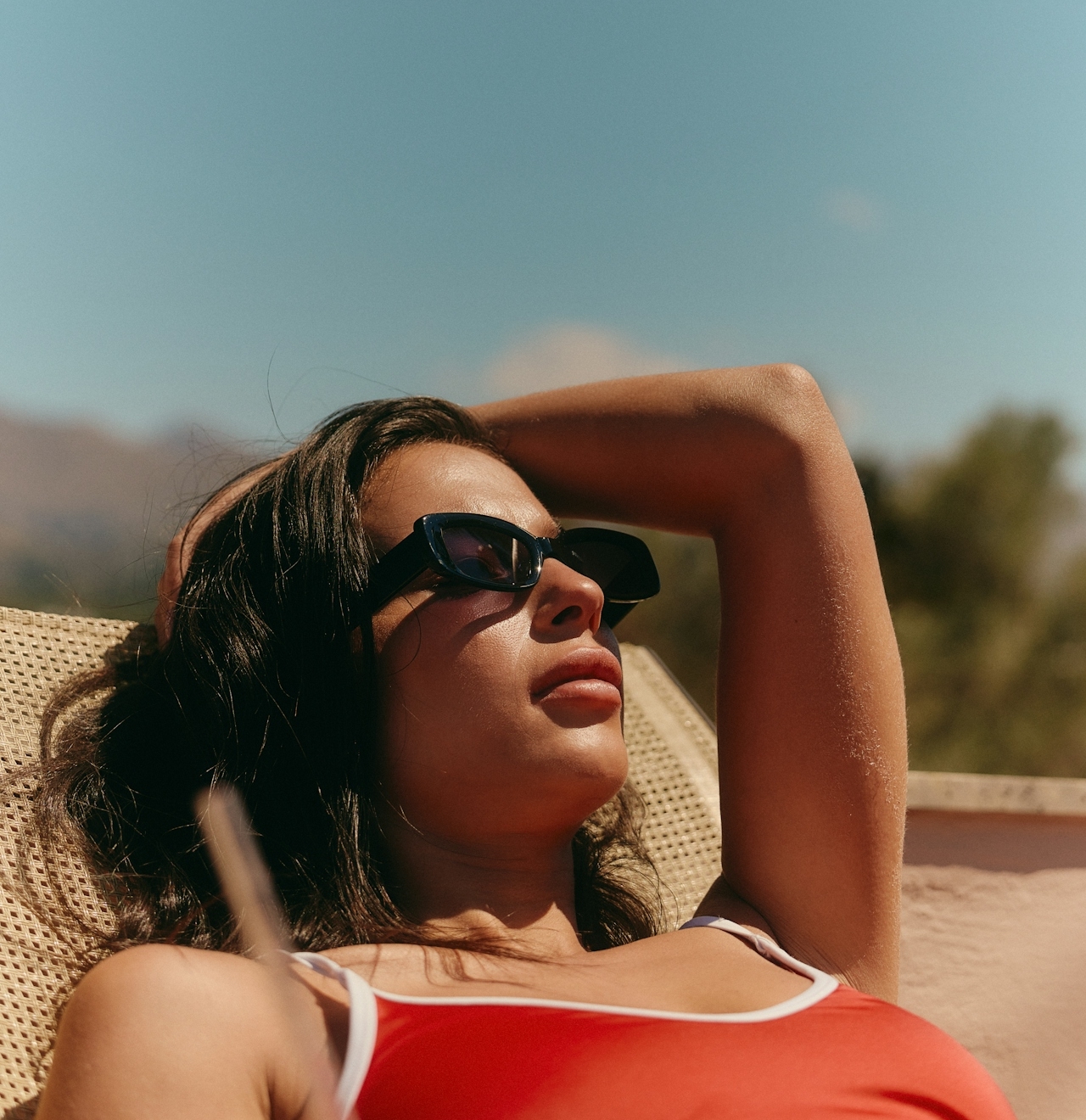A close-up of a woman sunbathing on a lounge chair wearing dark sunglasses and a red swimsuit against a clear blue sky.