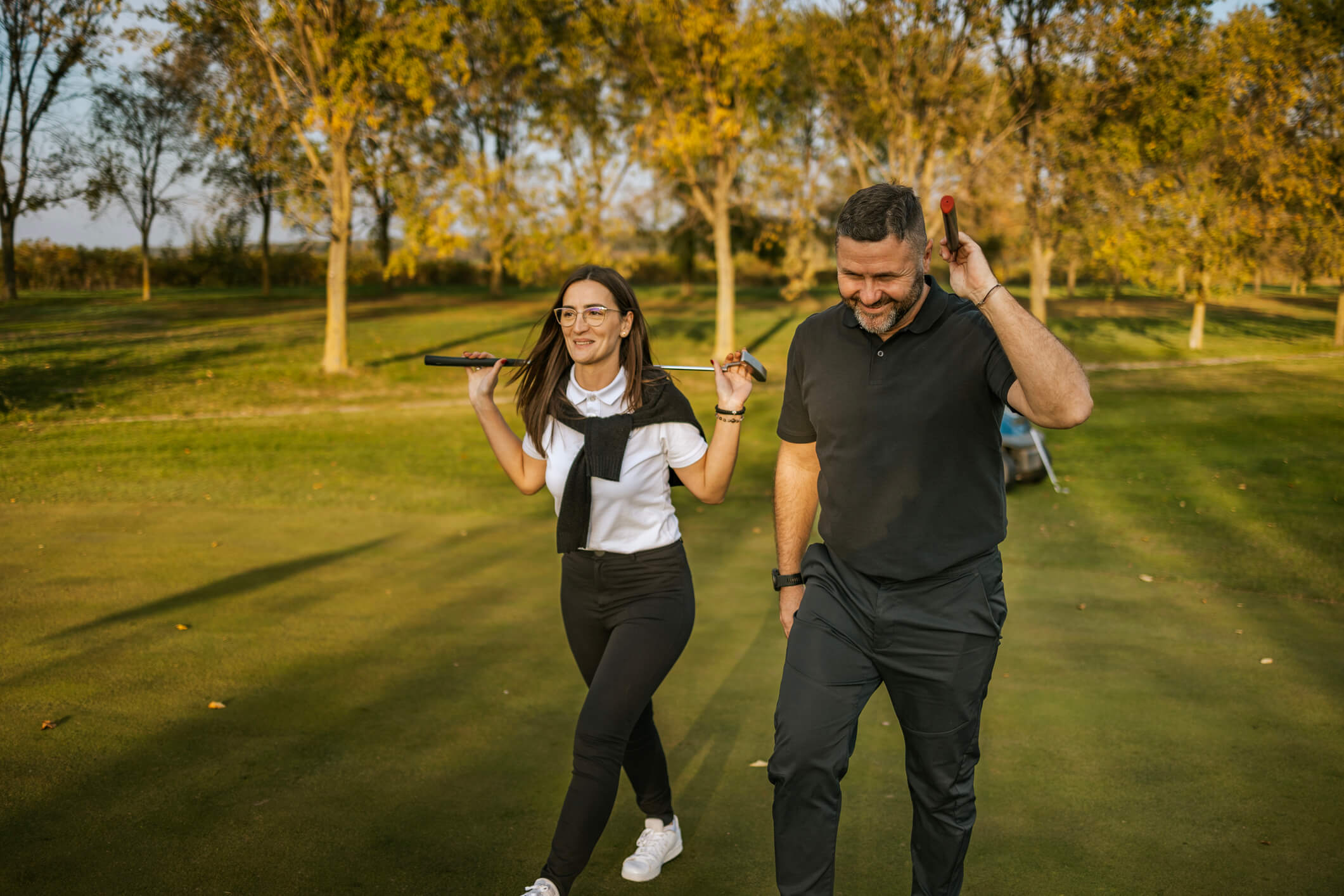 A smiling couple walking along a green golf course carrying clubs over their shoulders, with autumn trees in the background.