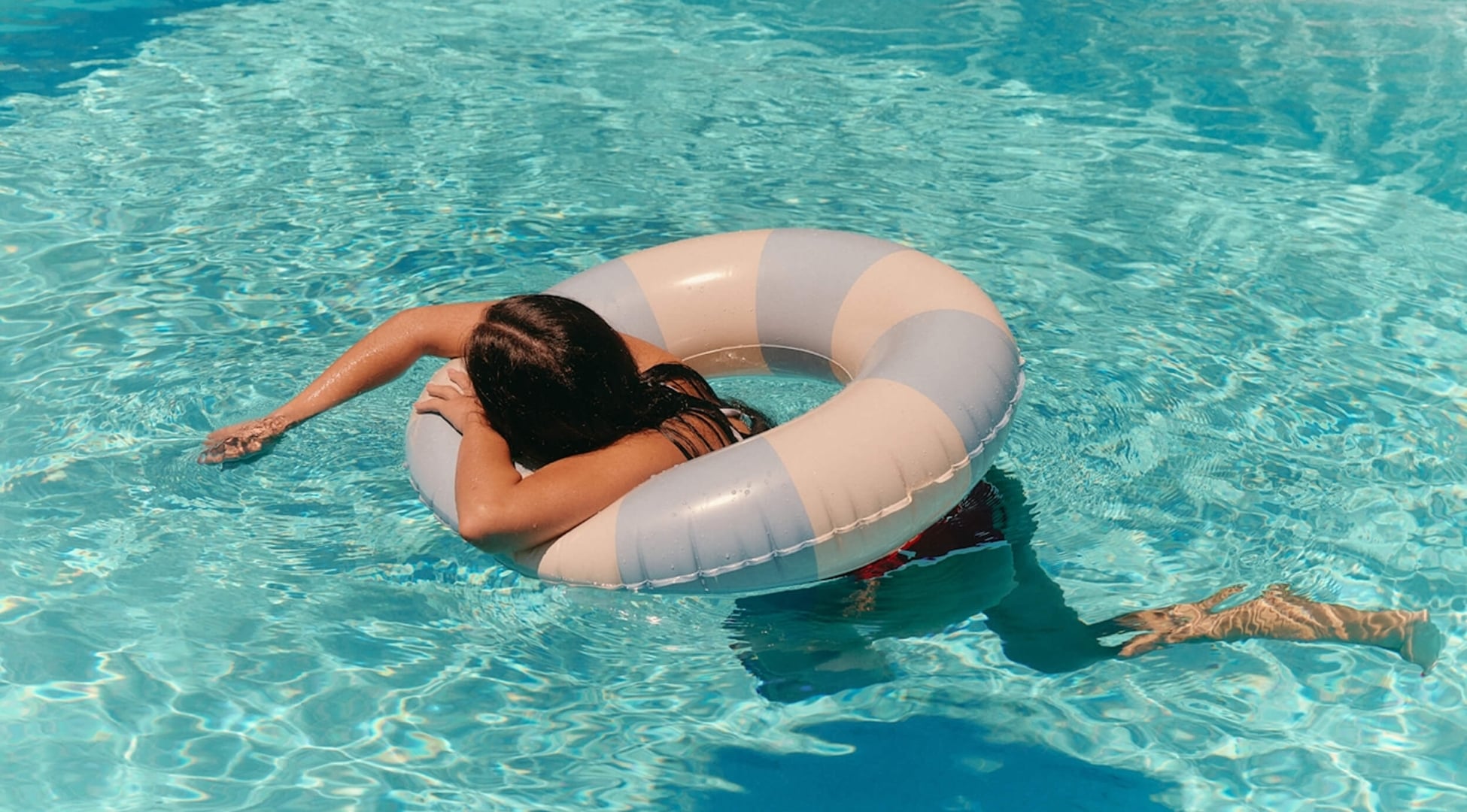 A person relaxing in a swimming pool, floating with their arms resting over a blue and white striped inner tube in crystal clear water.