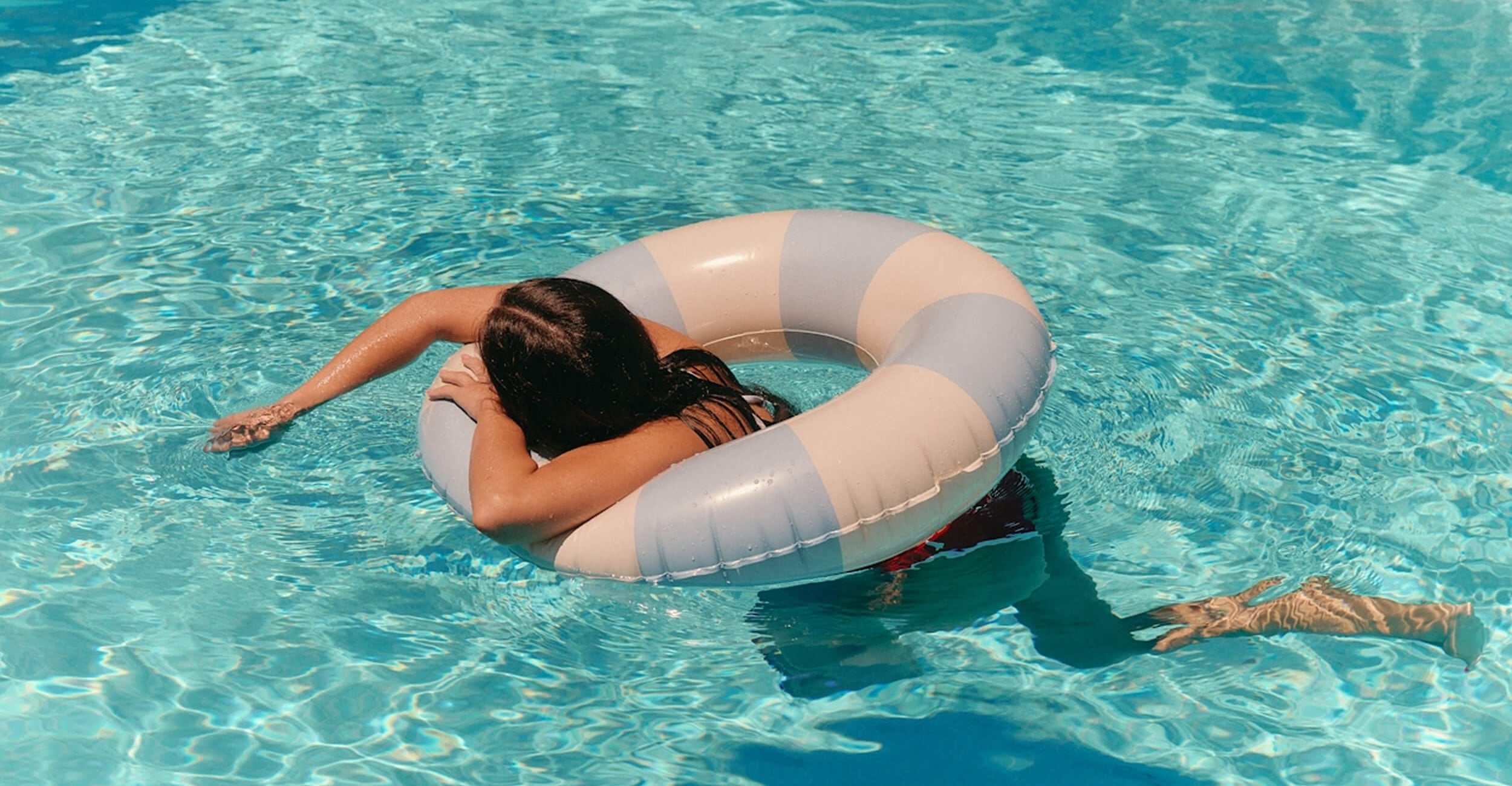 A person relaxing in a swimming pool, floating with their arms resting over a blue and white striped inner tube in crystal clear water.