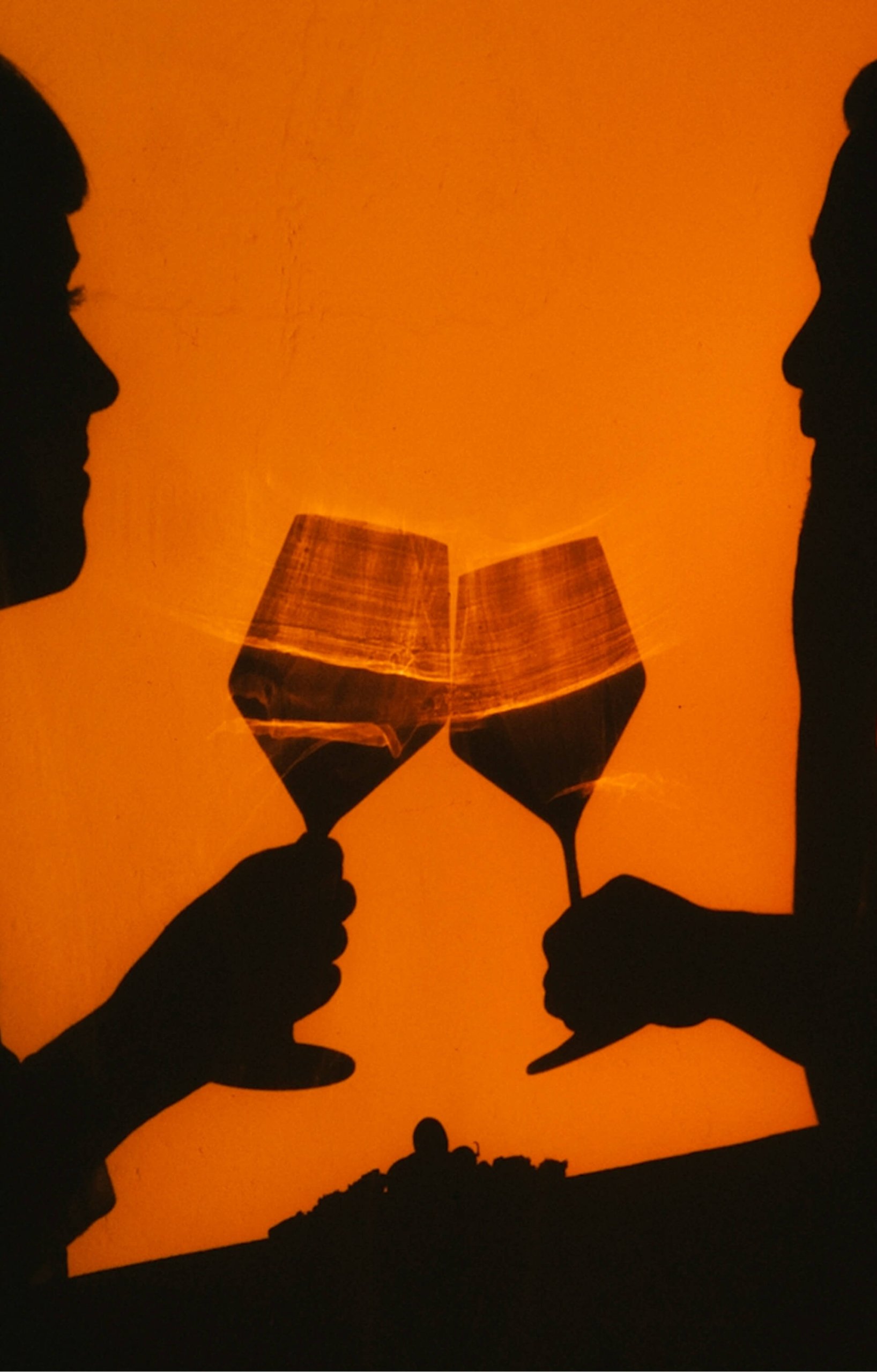 A silhouette of two people toasting with wine glasses against a glowing orange background, highlighting the liquid in the glasses.