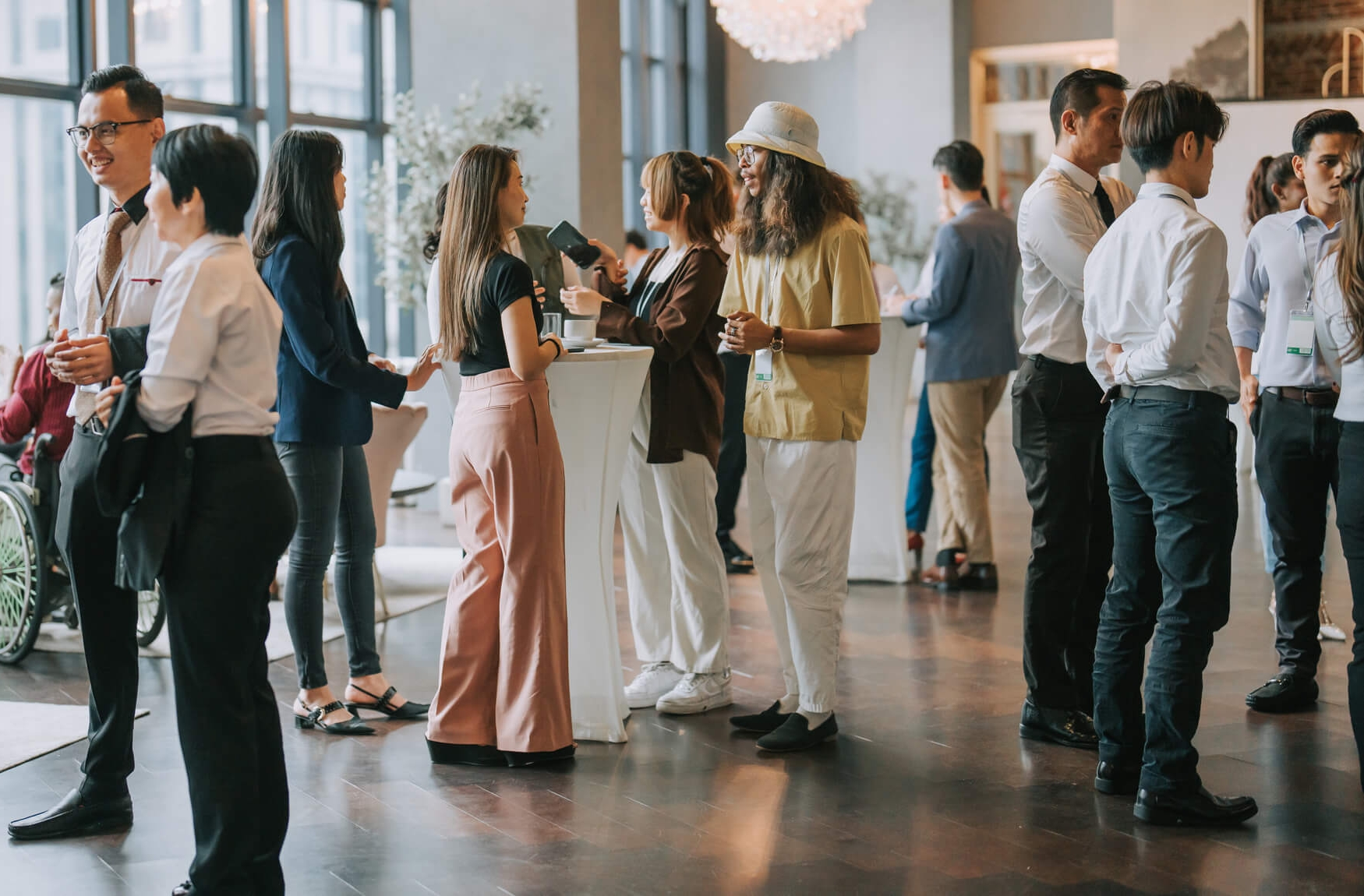A diverse group of professionals in business attire mingling and networking at a corporate event in a bright, modern venue with large windows.