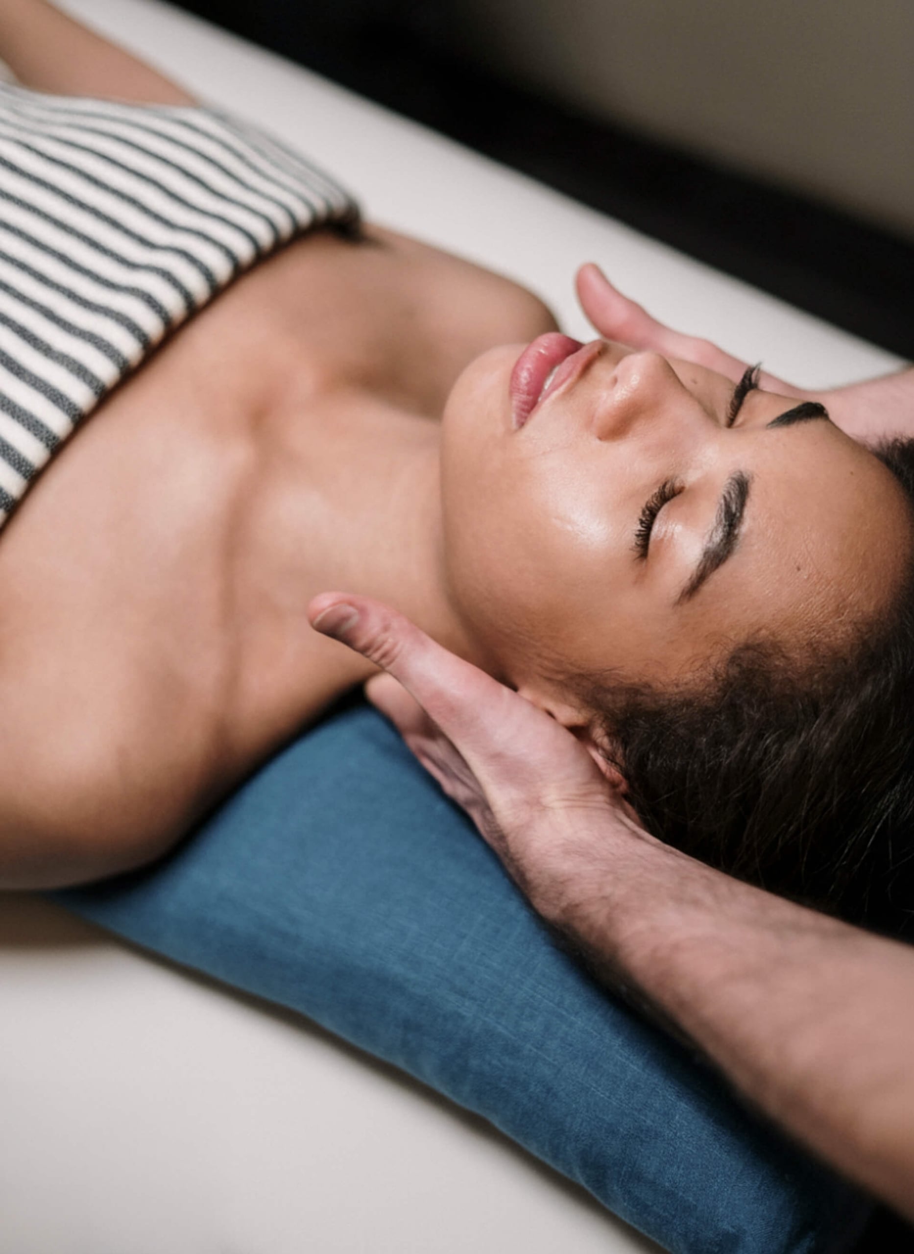 A close-up of a woman with her eyes closed receiving a relaxing neck and head massage from a therapist while lying on a blue pillow.