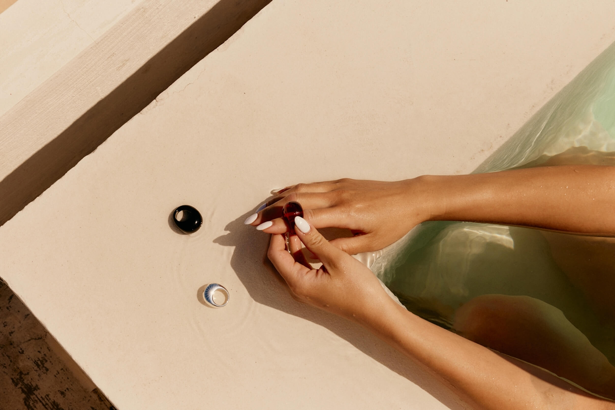 A pair of hands wearing rings resting on the beige edge of a pool or tub, partially submerged in clear, rippling water.