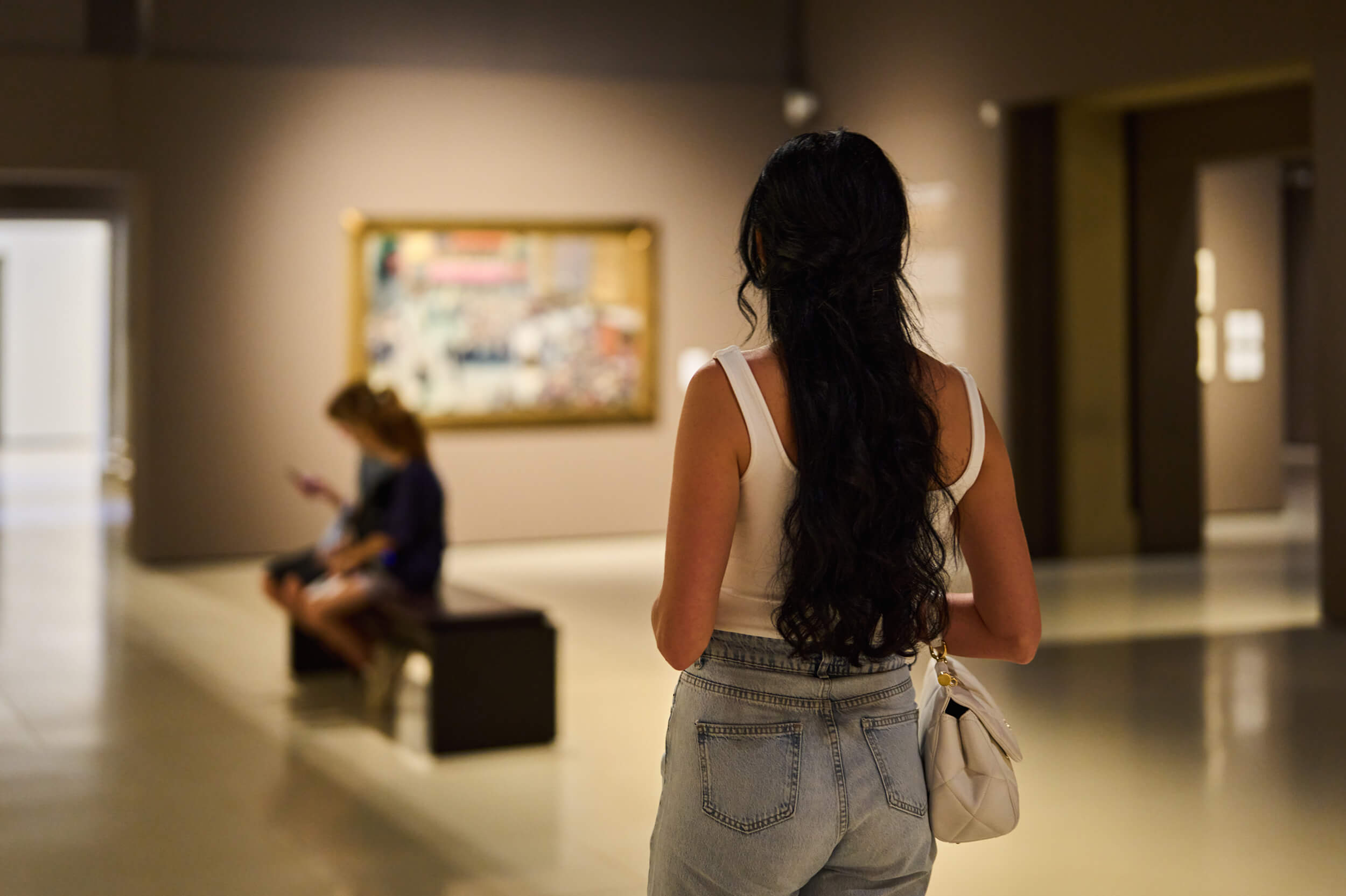 A woman with long dark hair viewing a framed painting in an art gallery, wearing a white top and jeans with a white purse.