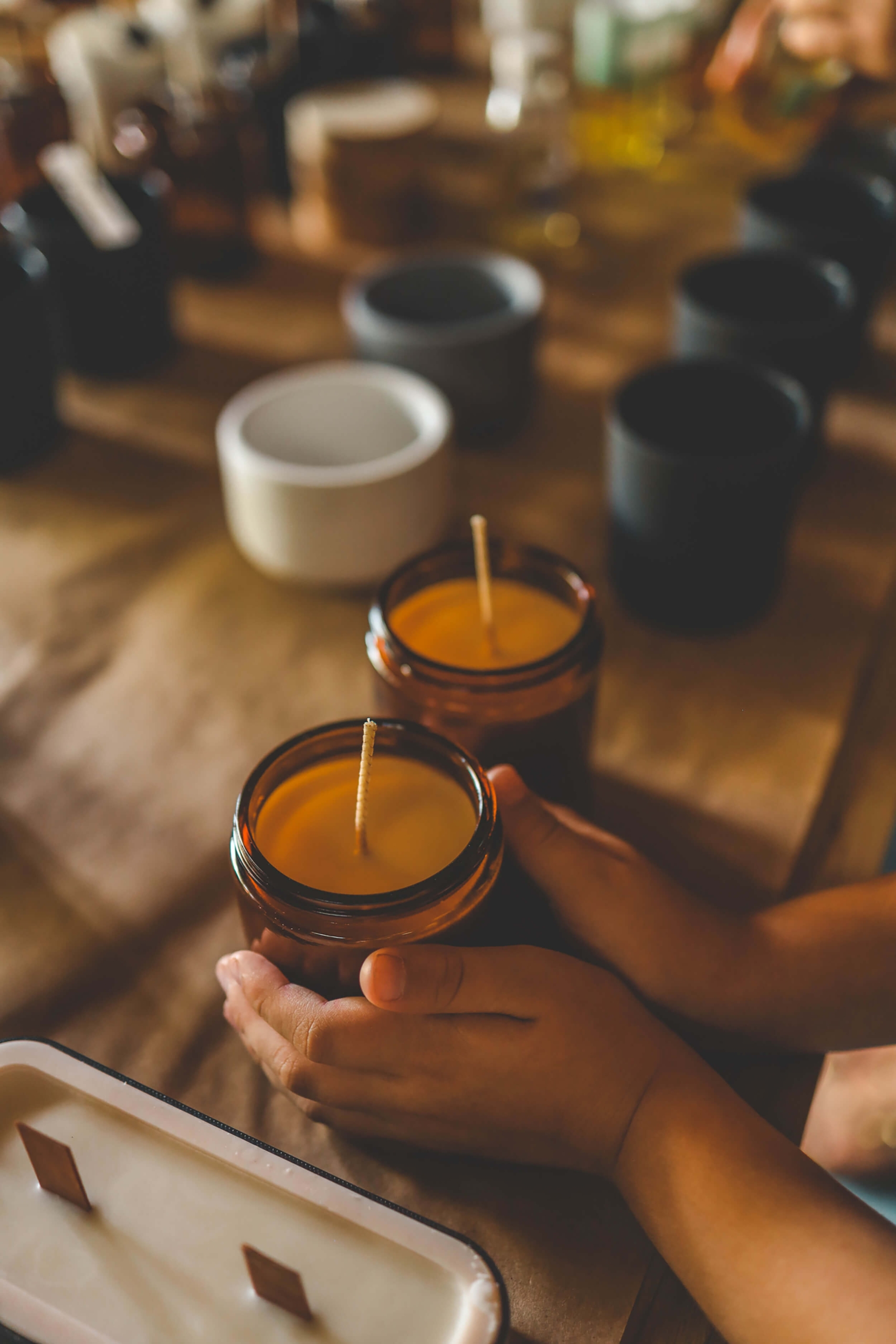 A pair of hands holding an amber glass candle jar with a wooden wick, surrounded by other candle containers on a table.