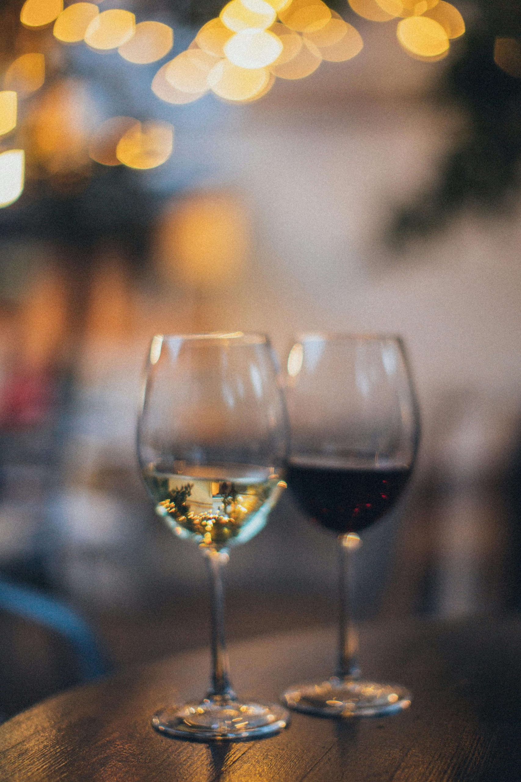 Two wine glasses, one filled with white wine and the other with red wine, sitting on a table with warm, blurred bokeh lights in the background.