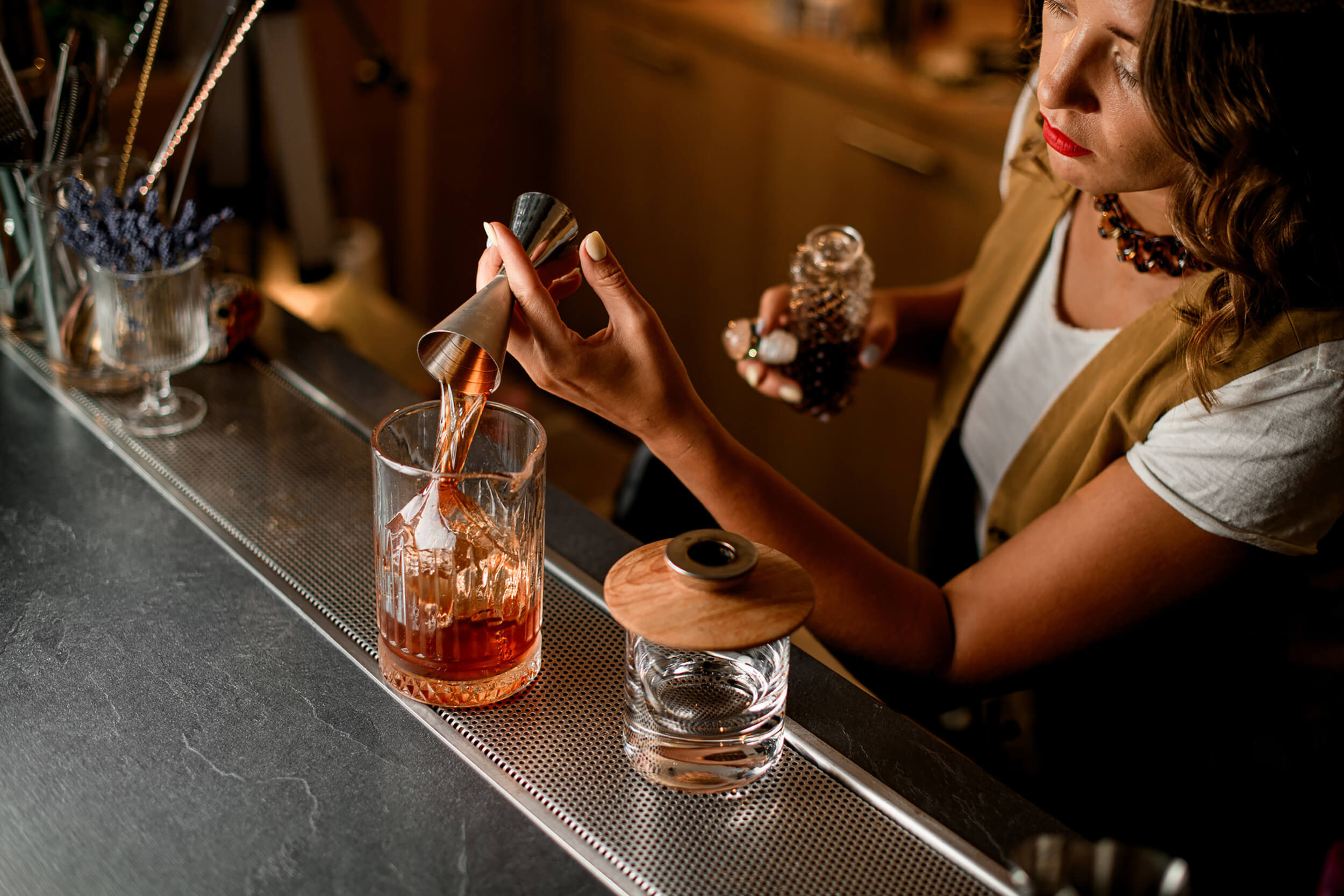A bartender pouring an amber cocktail from a metal jigger into a crystal glass containing a large ice cube.