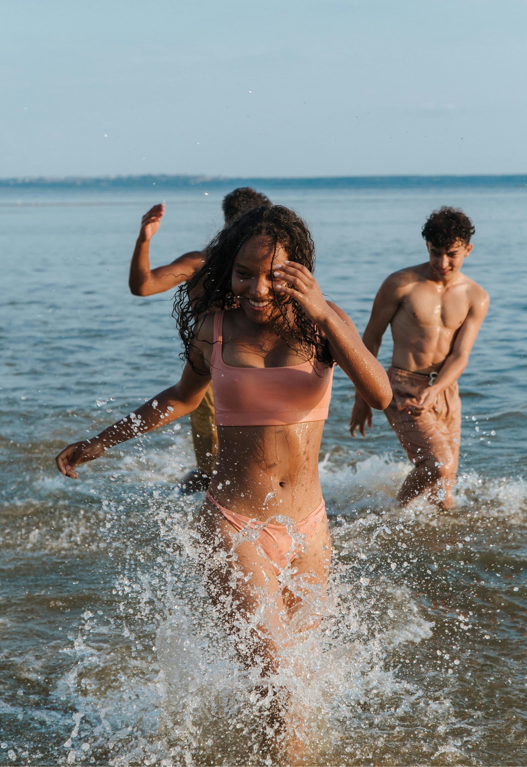 A group of young friends laughing and running through shallow ocean water, focusing on a smiling woman in a peach bikini splashing toward the camera.