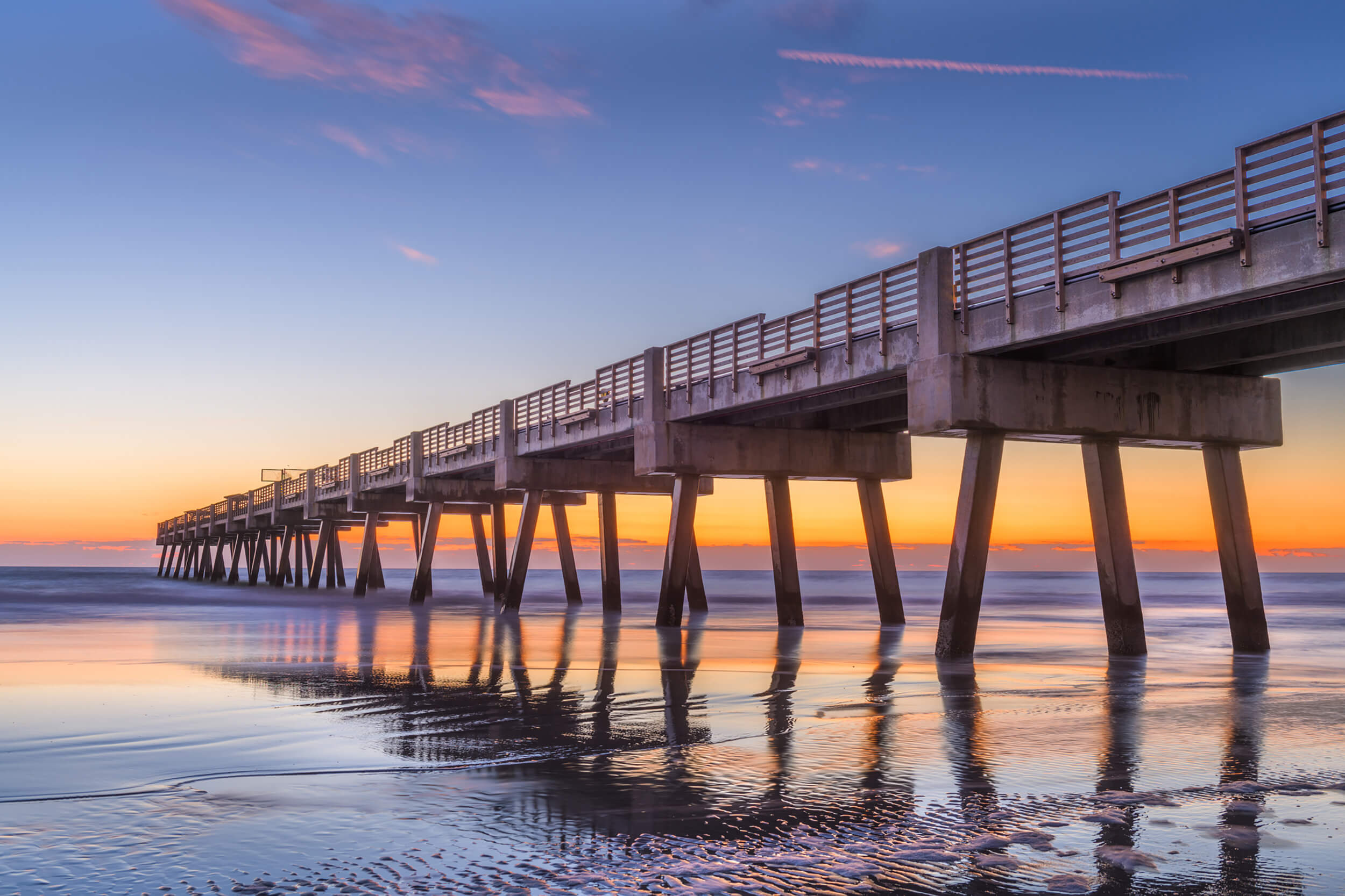 A view from the beach looking up at a long pier extending into the ocean at twilight, with pink and blue hues in the sky reflecting on the wet sand.