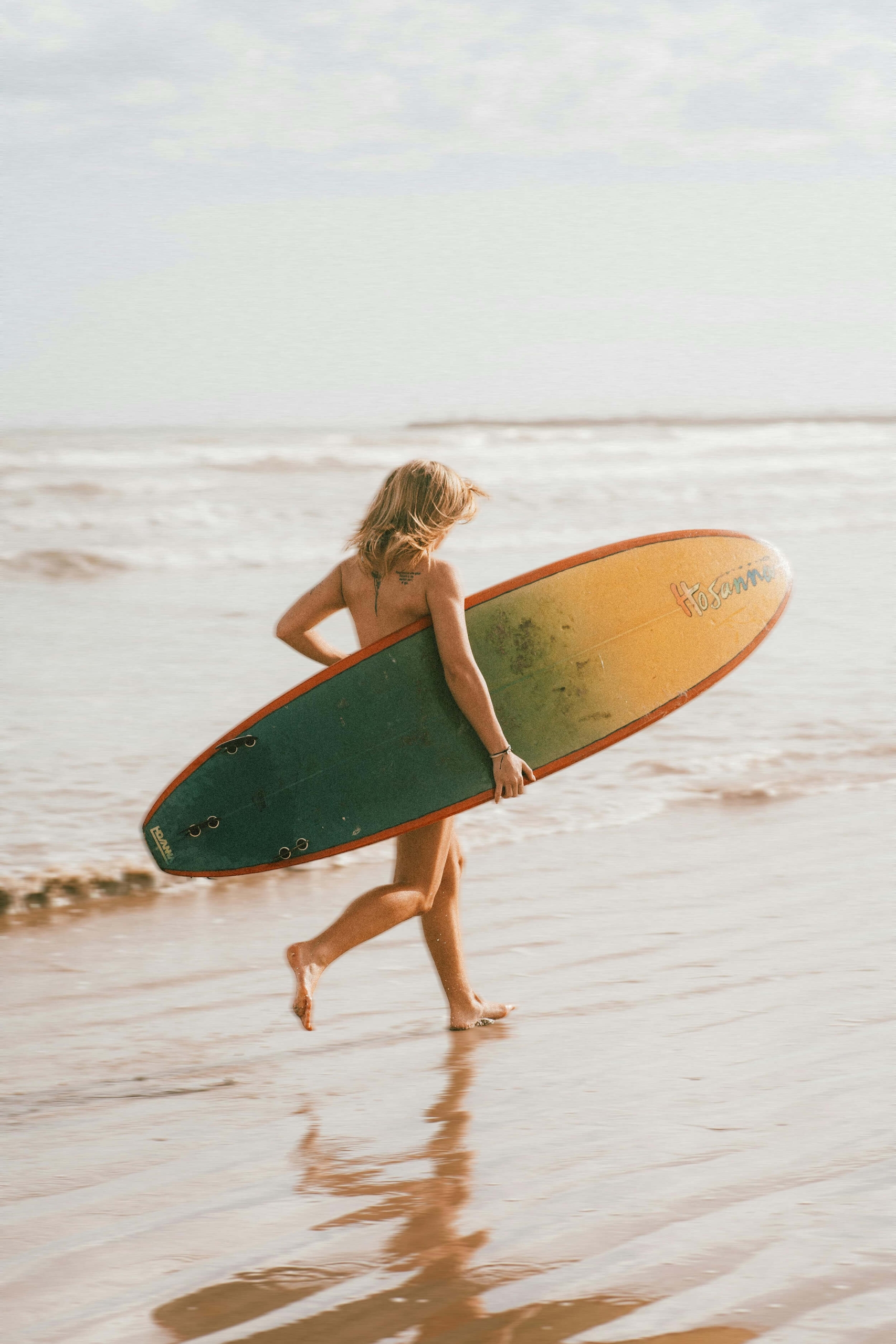 A surfer walking along the wet shoreline carrying a longboard with a yellow and green gradient design under their arm.