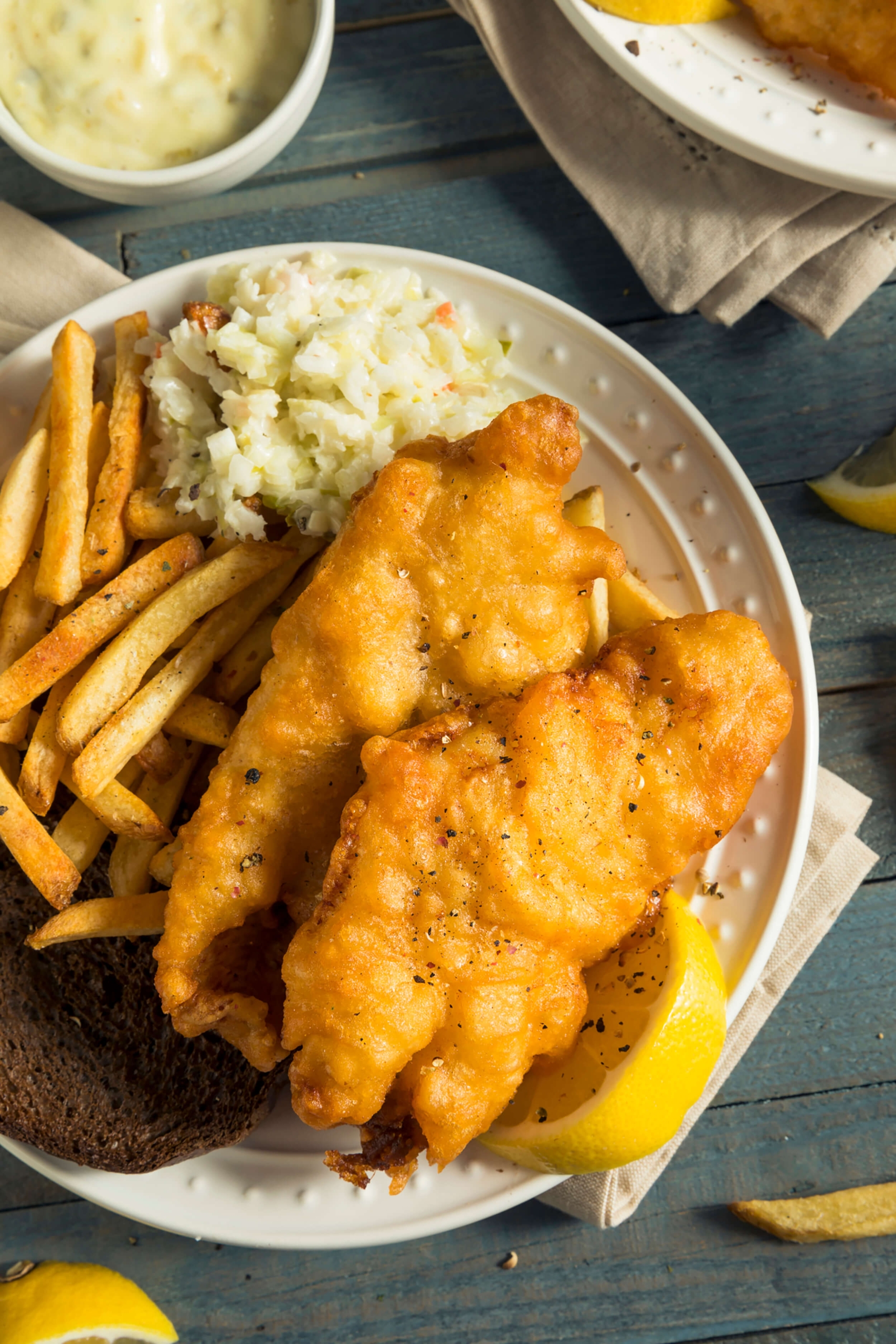 A plate of golden fried fish and french fries served with coleslaw and a lemon wedge, accompanied by a side of tartar sauce.