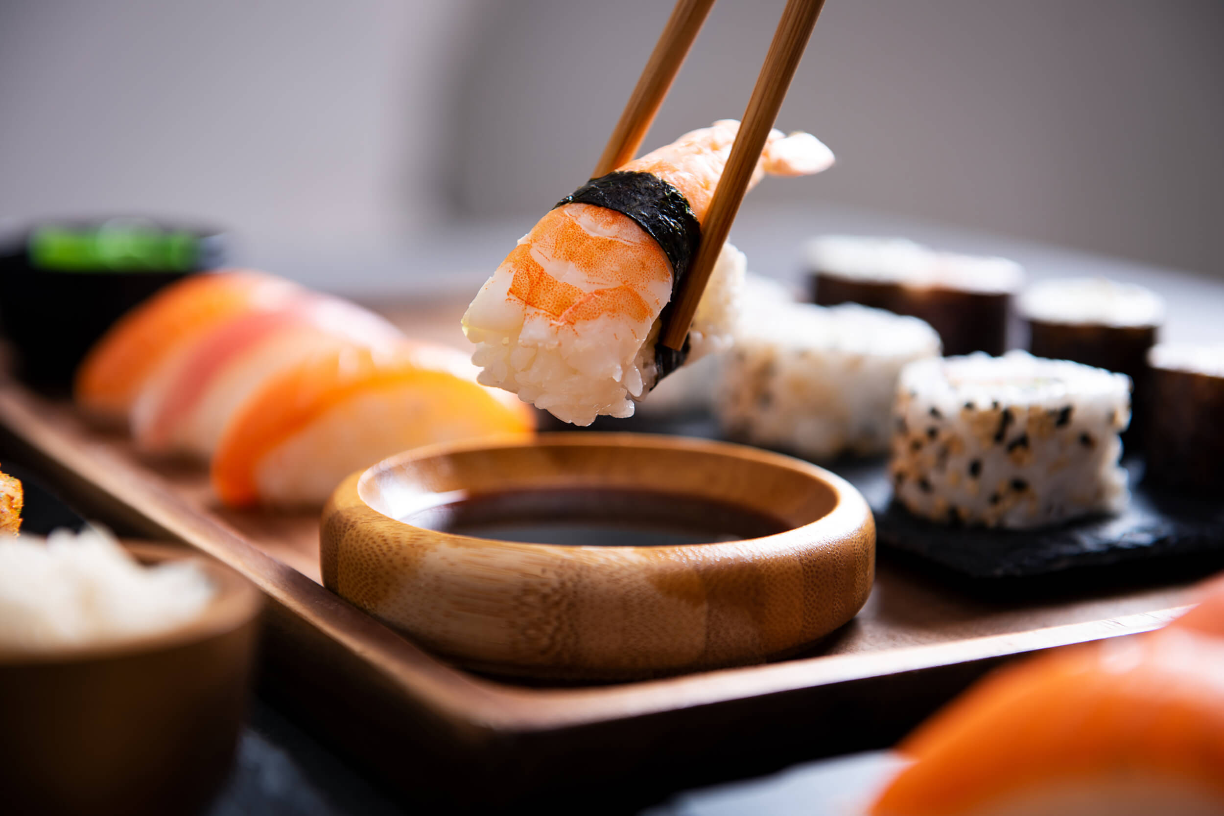 Wooden chopsticks holding a piece of shrimp nigiri over a small bowl of soy sauce, with a platter of sushi rolls in the background.