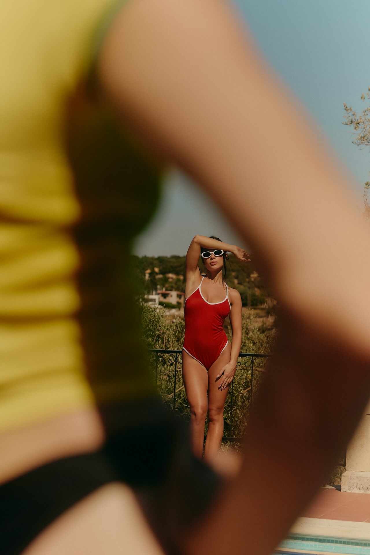 A woman in a red one-piece swimsuit shielding her eyes from the sun, framed artistically by an out-of-focus person in the foreground.