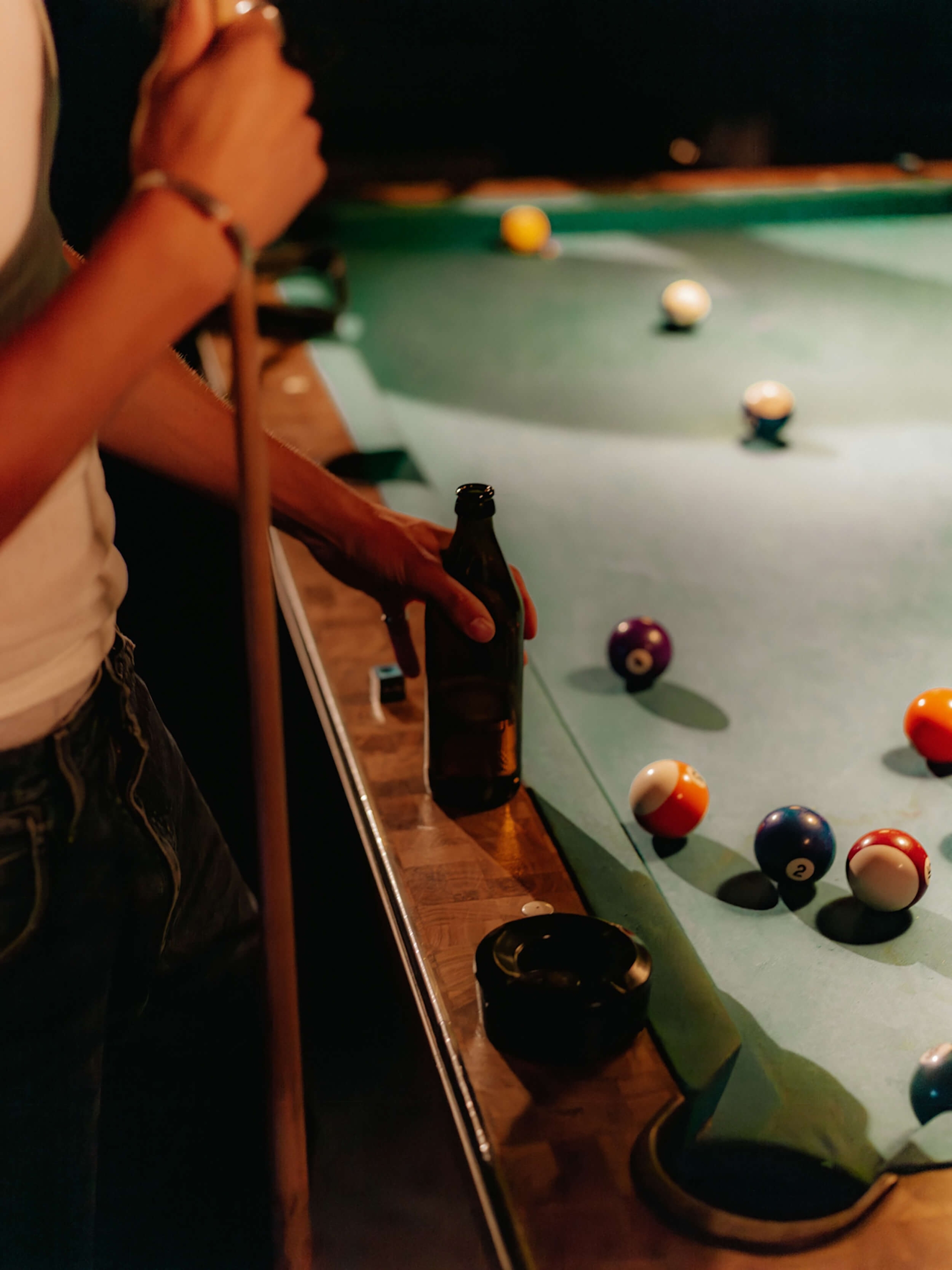 A close-up of a person holding a brown beer bottle on the wooden rail of a pool table, with scattered billiard balls on the green felt surface.
