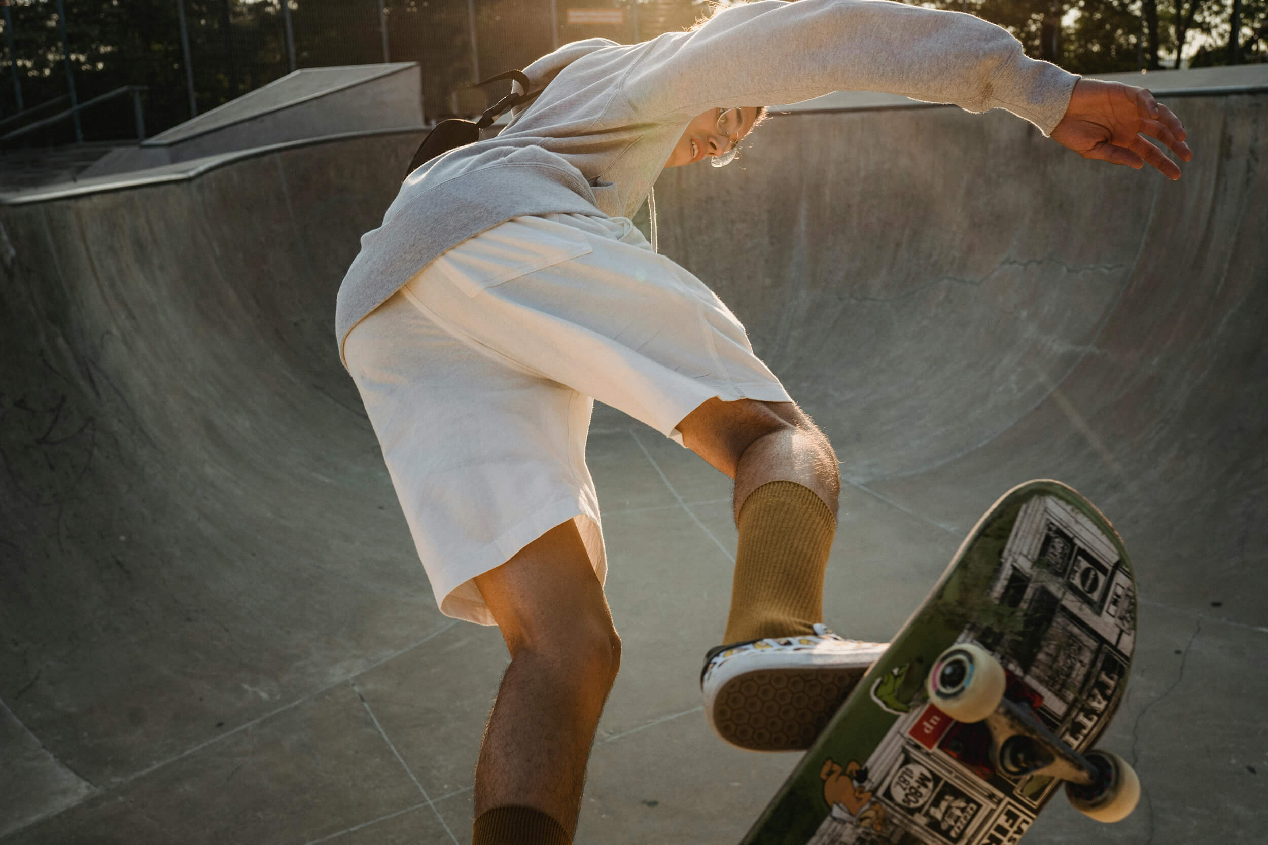 A skateboarder performing a mid-air trick in a concrete bowl at a skate park, illuminated by a bright sun flare.