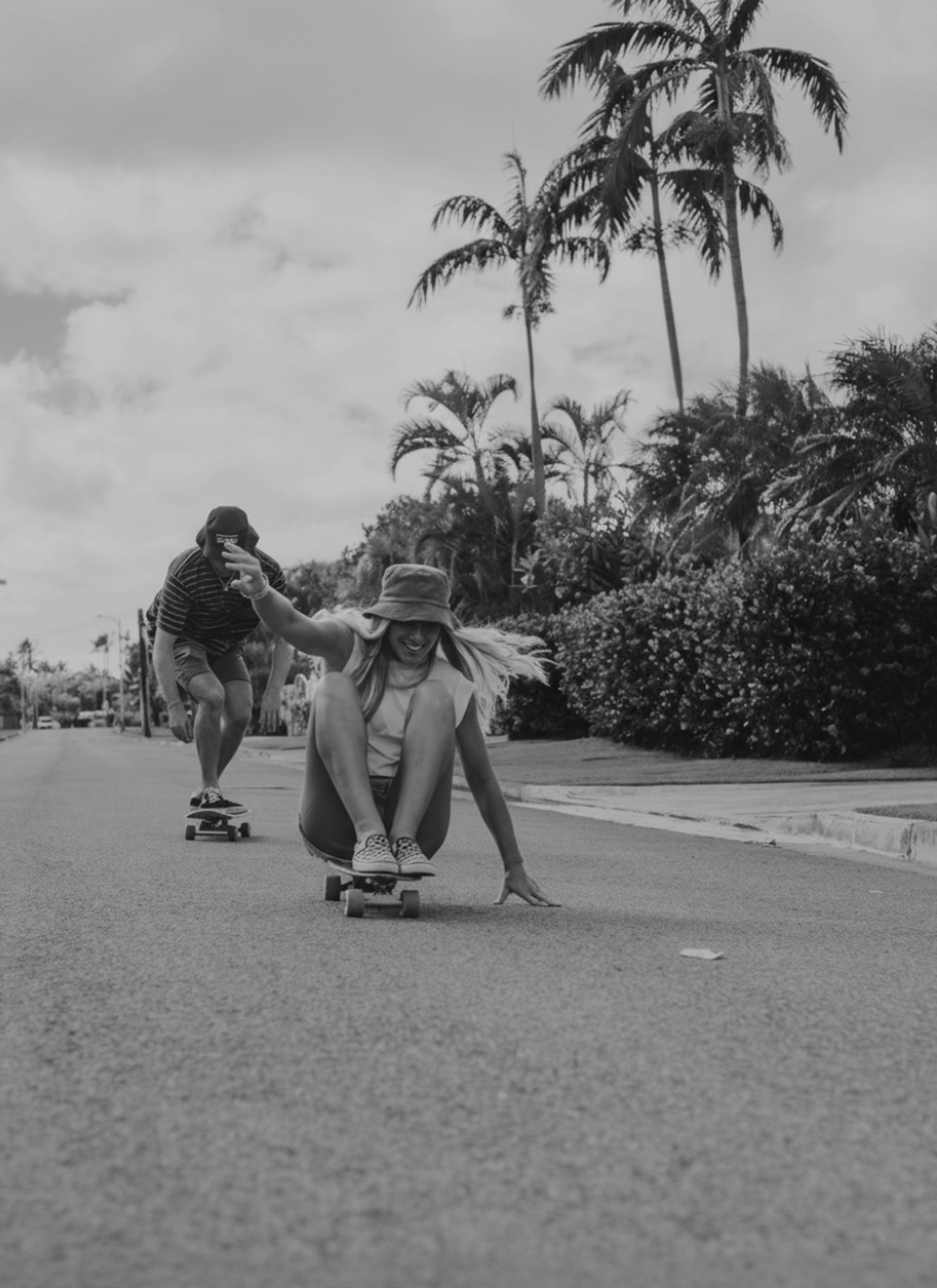 A black-and-white photo of a couple skateboarding down a palm tree-lined street; the woman sits on her board while the man skates behind her.