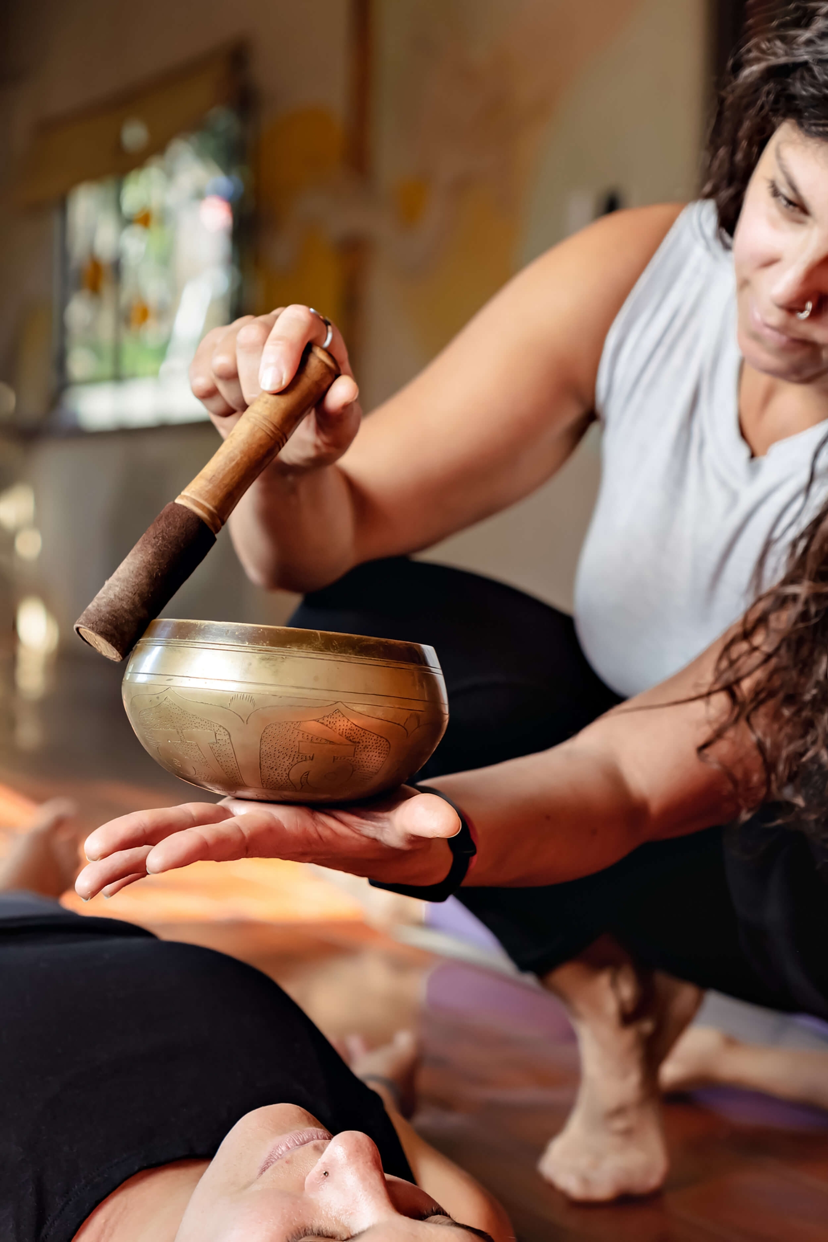 A sound healer holding a brass singing bowl and striking it with a mallet over a client lying down during a therapy session.