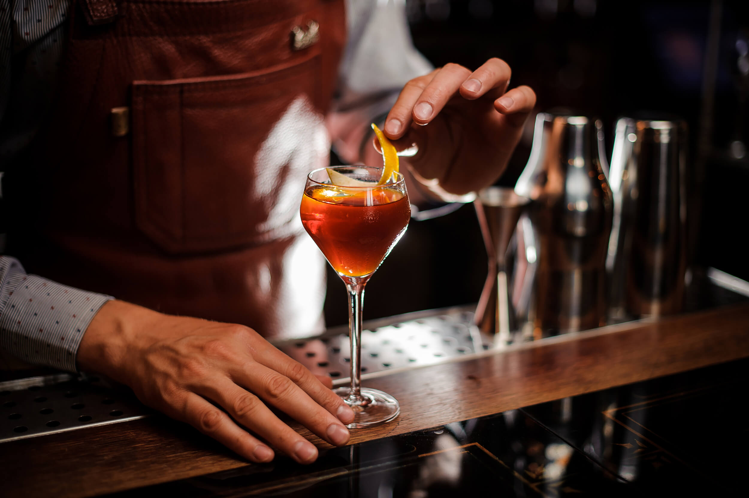 A bartender wearing a leather apron carefully placing a lemon peel garnish onto an amber-colored cocktail in a stemmed glass.