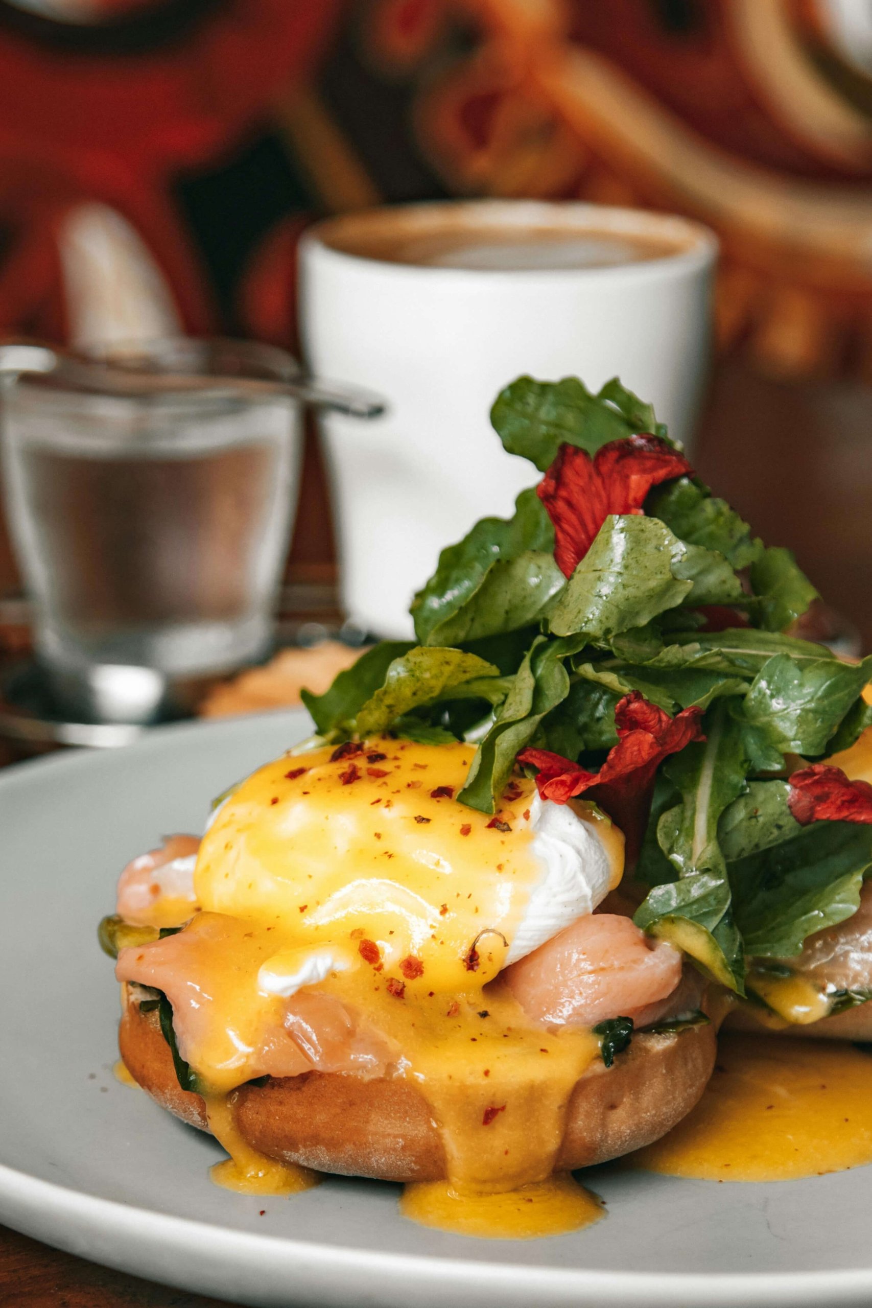 Close-up of a biscuit breakfast sandwich topped with smoked salmon, a poached egg, hollandaise sauce, and chili flakes, served with a side of greens and edible flowers.