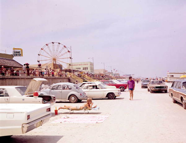 A vintage photograph of a busy beach day showing rows of classic cars parked on the sand near a boardwalk and Ferris wheel.
