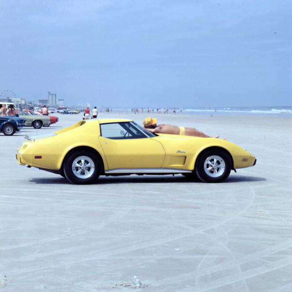 A bright yellow vintage sports car parked on the beach sands with a person resting on the hood, looking out at the water.