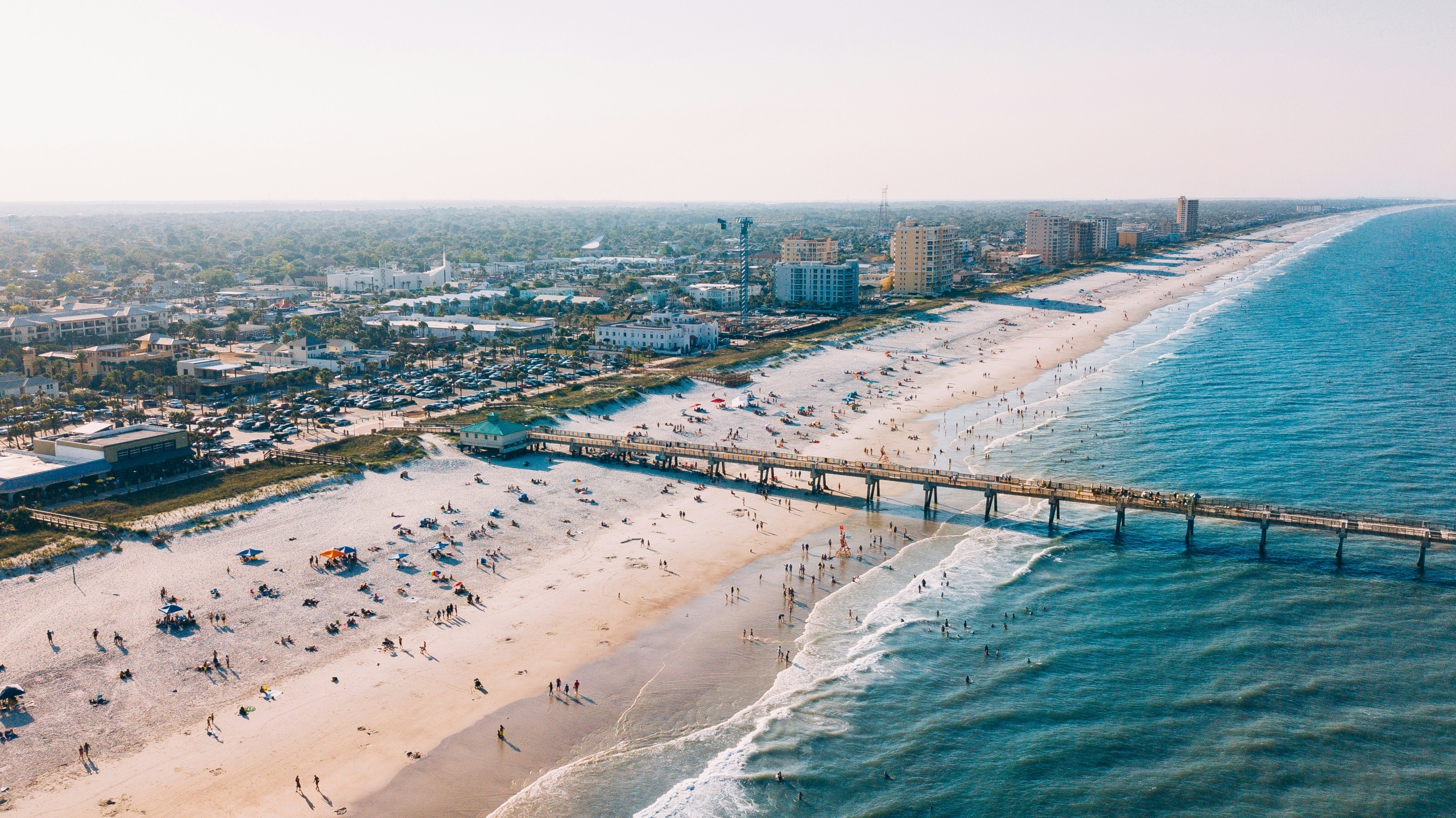 An aerial view of a long fishing pier extending into the blue ocean from a white sandy beach lined with buildings and palm trees.