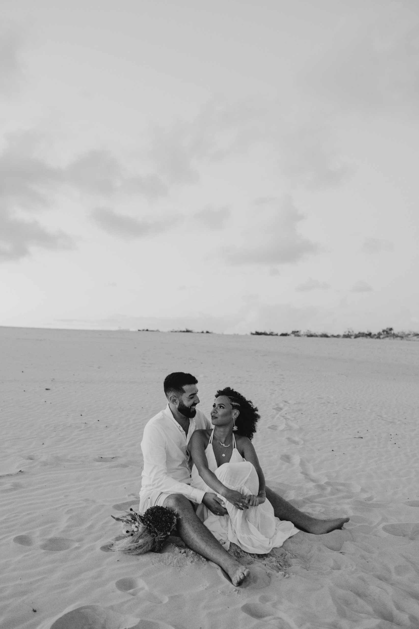 A couple dressed in white sitting together on sand dunes, smiling and looking at each other with a cloudy sky in the background.