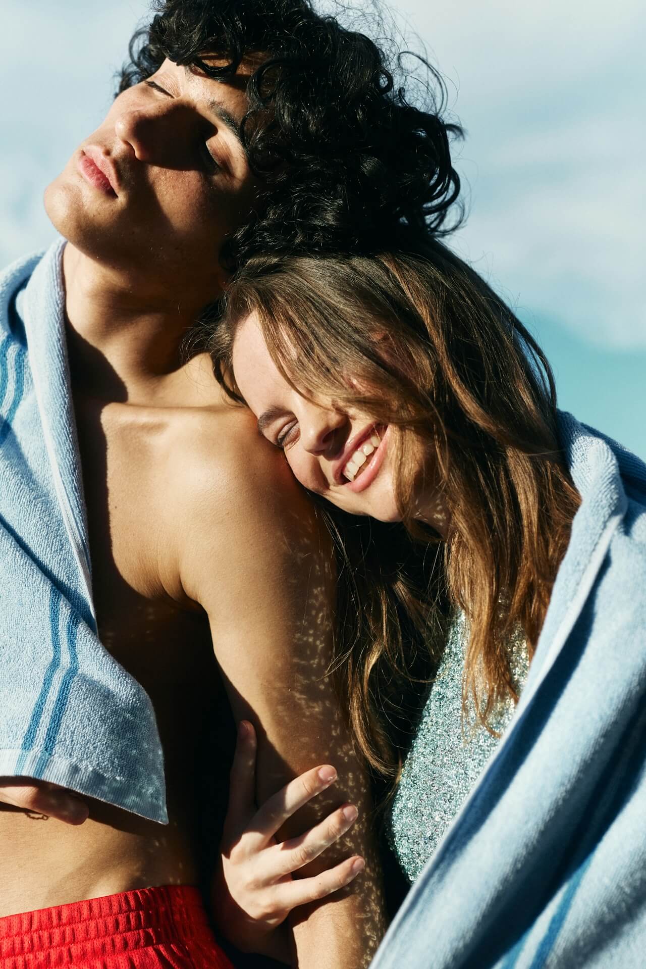 A close-up portrait of a young couple basking in the sun wrapped in light blue towels; the woman rests her head on the man's shoulder while smiling.