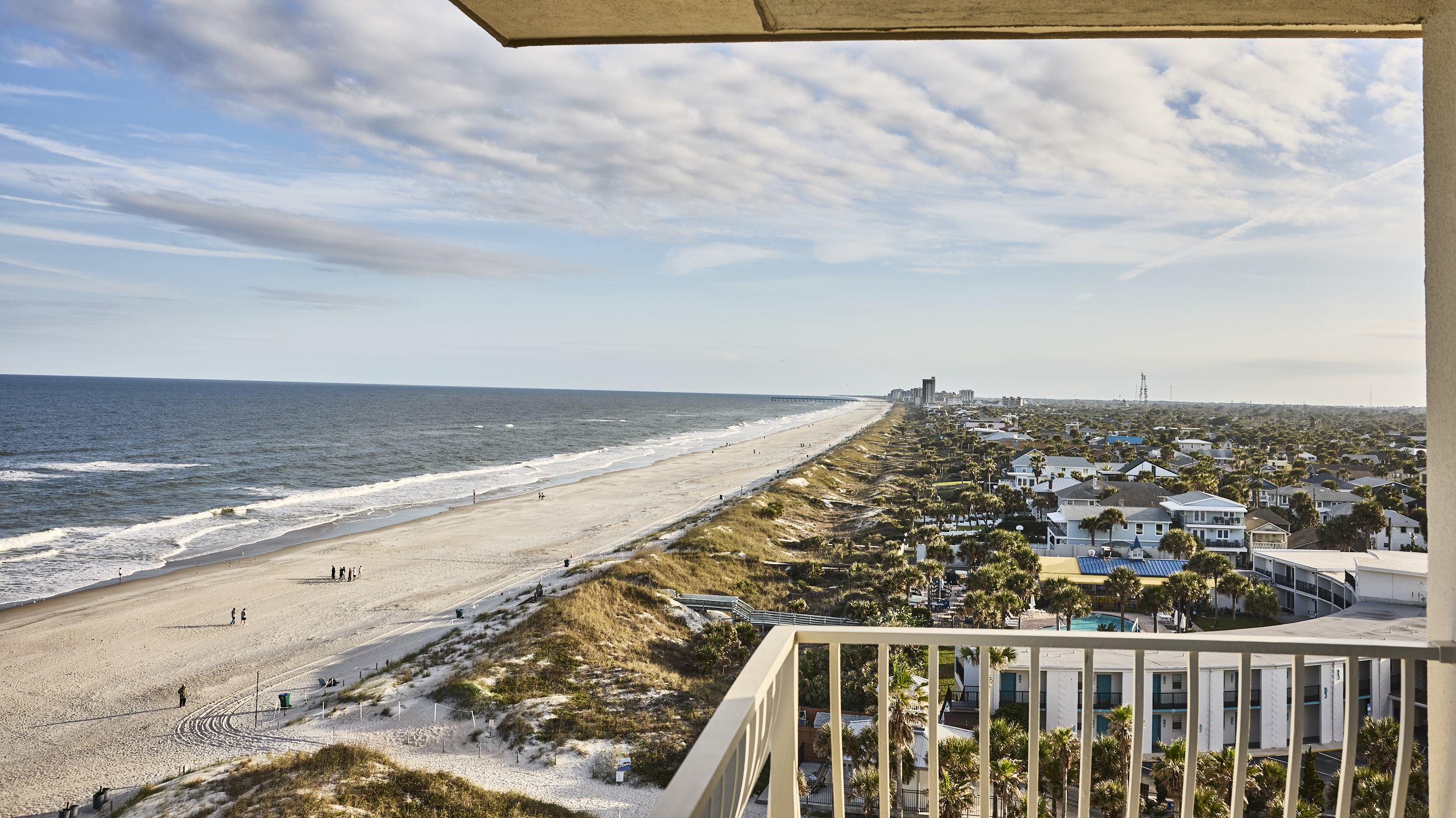 High-angle balcony view of a wide sandy beach, green dunes, and coastal buildings stretching into the distance.