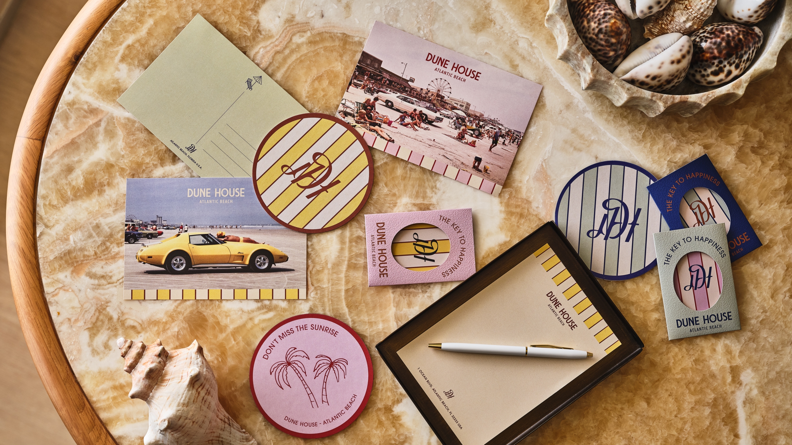 Overhead view of Dune House stationery, branded coasters, and a white pen on a marble tabletop with seashells.