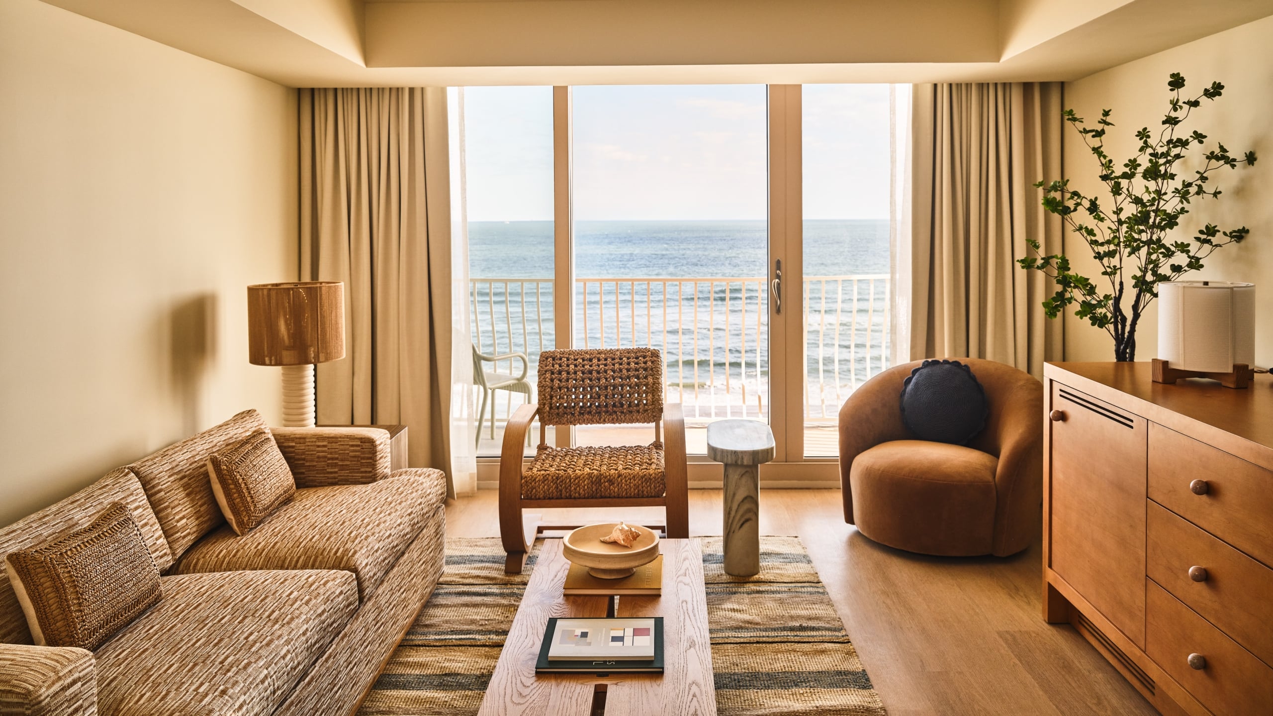 A hotel living area with a textured sofa, woven chair, and floor-to-ceiling glass doors opening to the beach.
