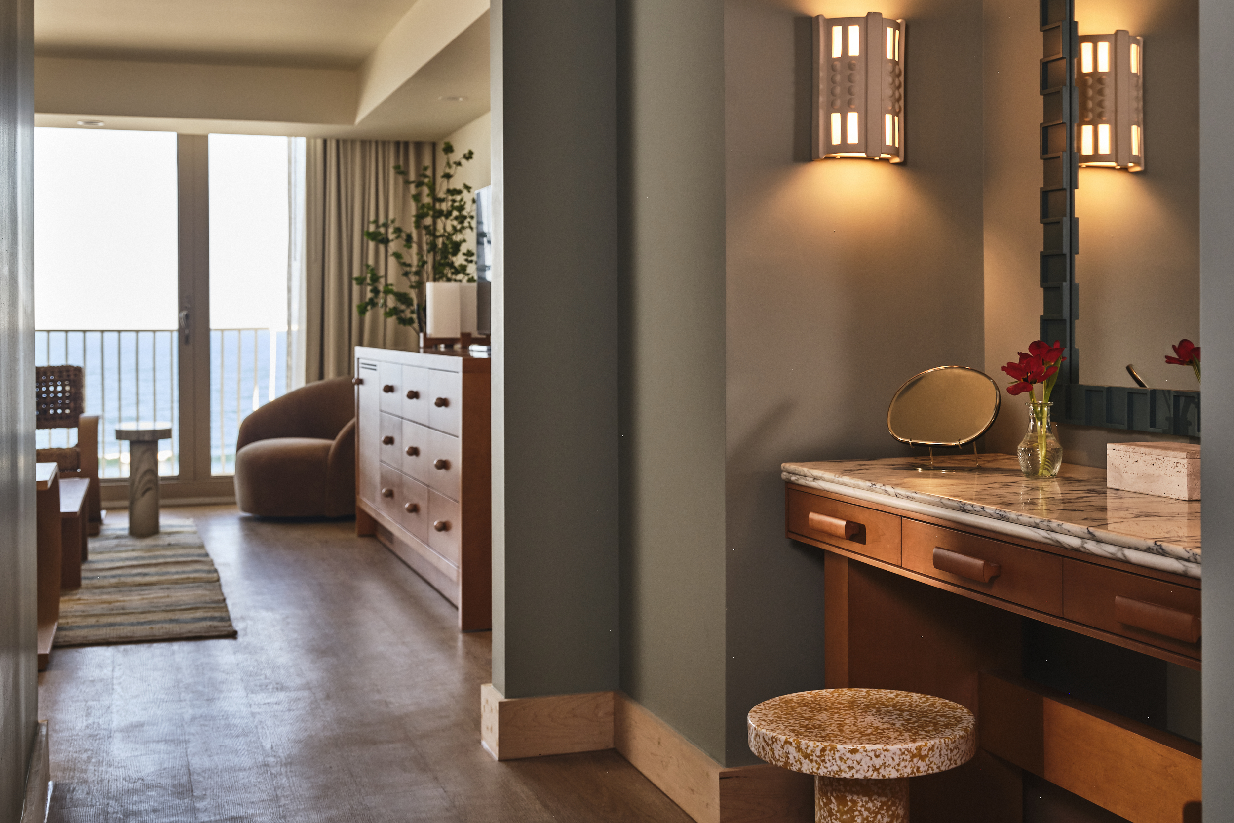 View from a hallway looking past a marble vanity into a sunlit hotel suite with a beachfront balcony.